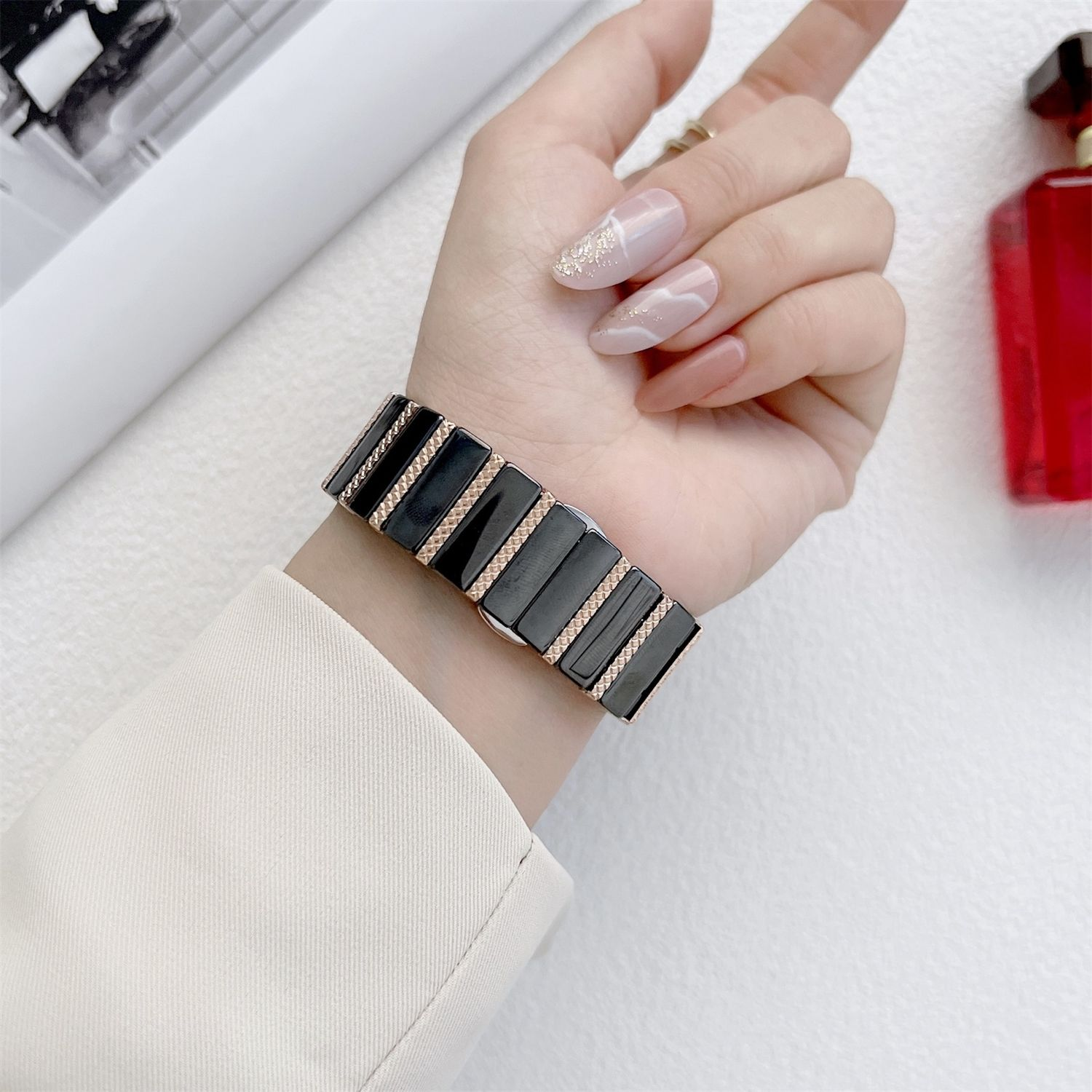 Eine Hand, die eine schwarz-goldene Uhr trägt. Die Nägel sind hellrosa und goldglitzernd lackiert. Eine weiße Jacke ist sichtbar.