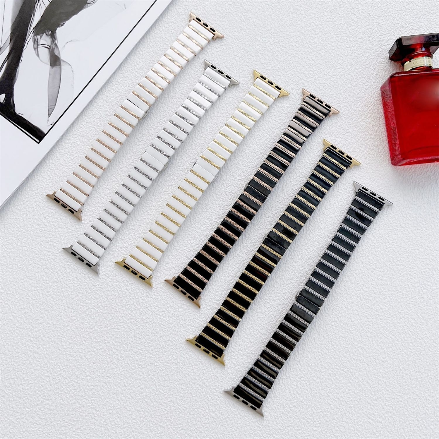 Uhrenarmbänder in verschiedenen Farben: Gold, Silber, Weiß und Schwarz, angeordnet auf einer weißen Oberfläche, mit einer roten Parfümflasche.