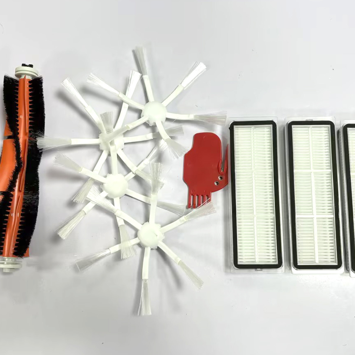 Części robota sprzątającego: wałek ze szczotką, białe szczotki, czerwone narzędzie i filtry, ułożone na białej powierzchni.