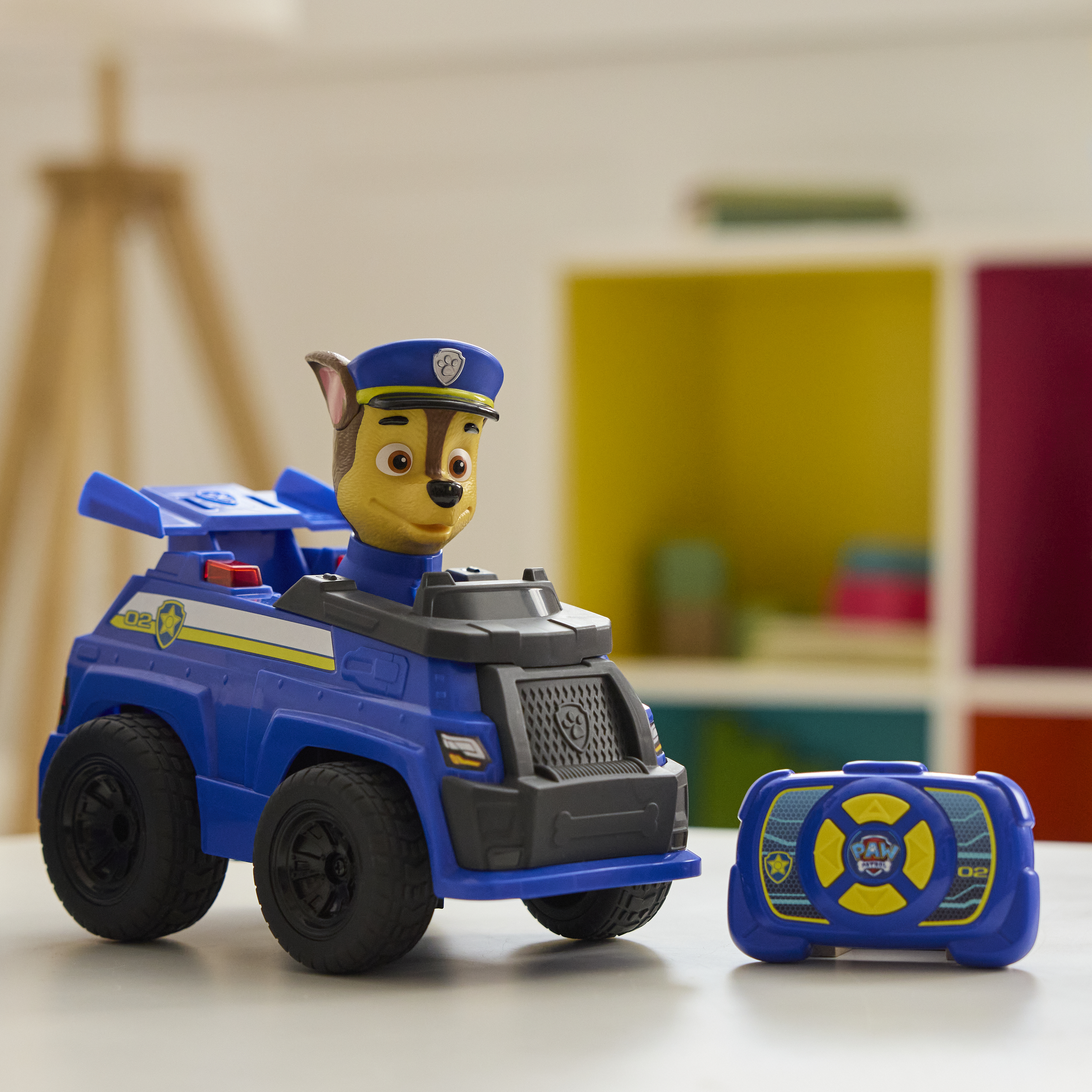 Niebieski samochód zabawkowy Paw Patrol z figurką Chase'a. Obok samochodu znajduje się niebieski pilot. Tło ma białe i kolorowe bloki.