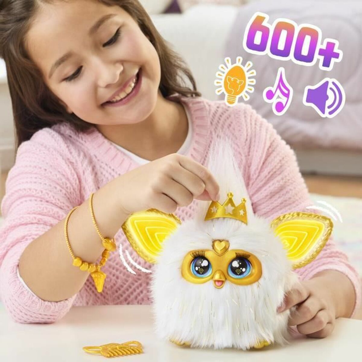 Ein Mädchen lächelt und hält ein weißes und goldenes Spielzeug. Das Spielzeug hat große Augen und eine Krone. Auf dem Tisch liegen ein goldenes Armband und ein Kamm.