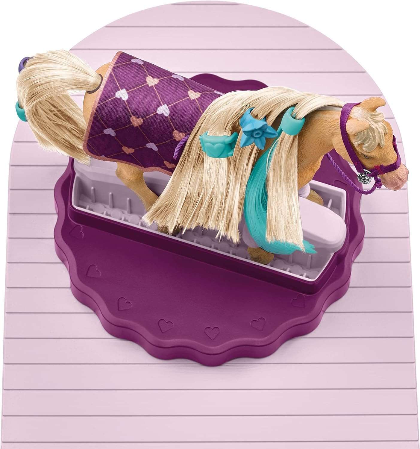Ein Spielzeugpferd mit blondem Haar, verziert mit blauen Accessoires, trägt eine lila Decke mit Herzmustern und liegt in einer lila Schale.