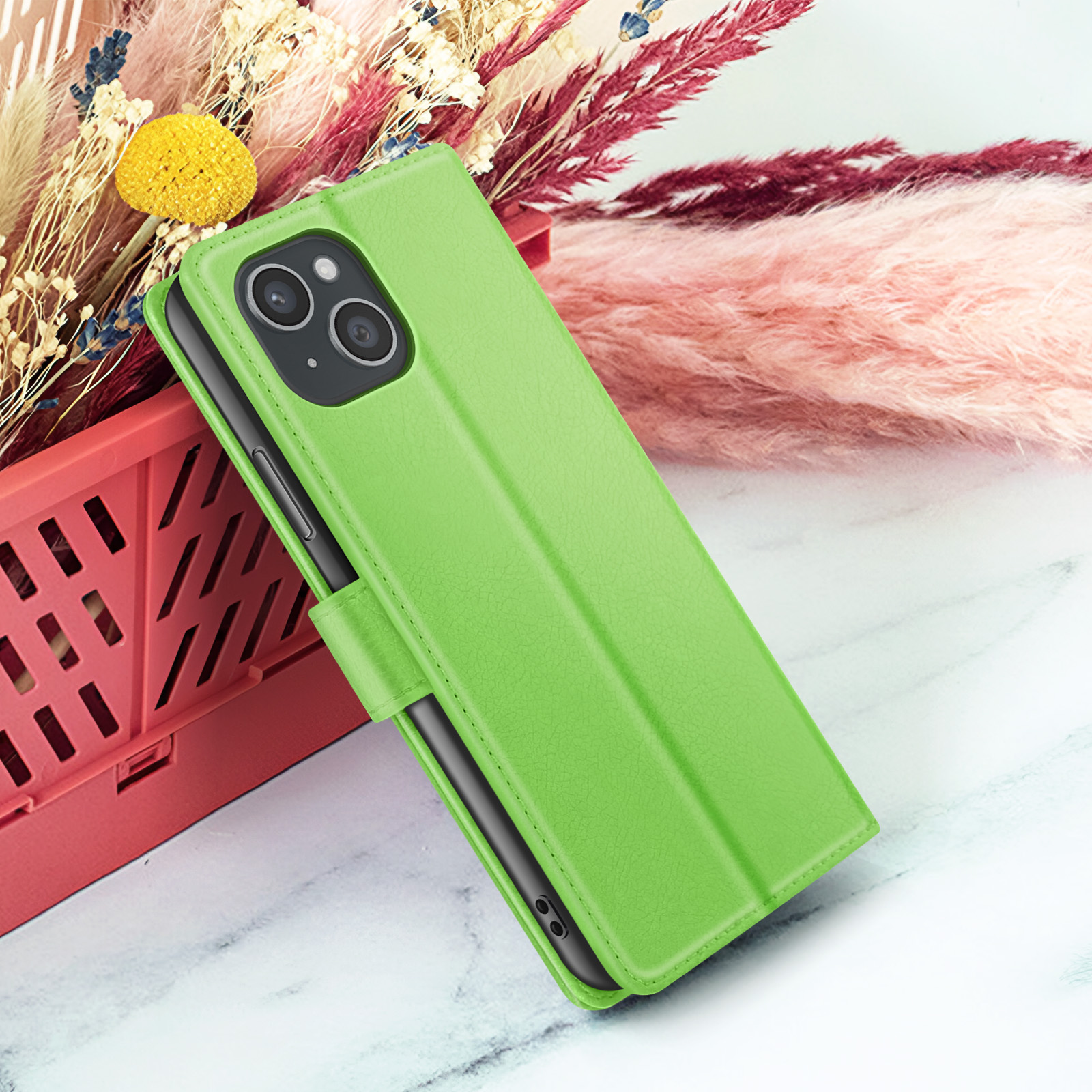 Een groene telefoonhoes met een telefoon erin, geplaatst voor roze en rode bloemen. De achtergrond is een marmeren oppervlak.