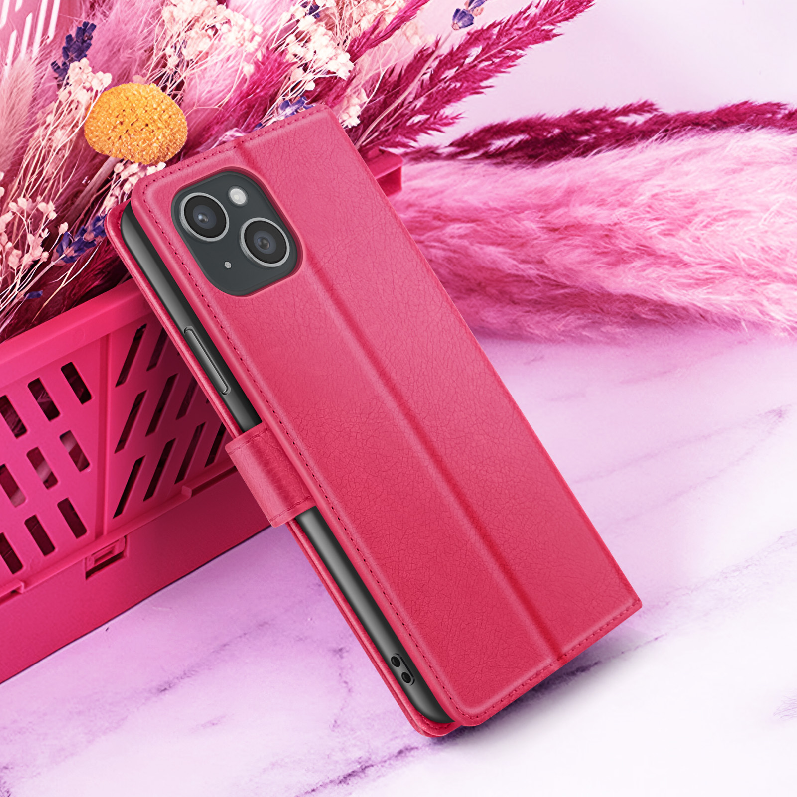 Een roze telefoonhoesje met een telefoon erin, geplaatst voor roze bloemen en een roze mandje op een marmeren oppervlak.