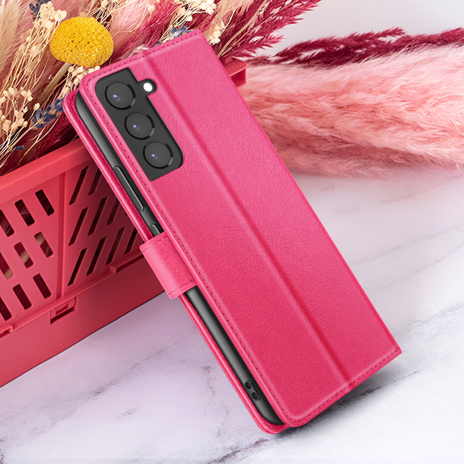Un étui de téléphone rose est ouvert. Il est à côté d'un panier rouge et de fleurs roses sur une surface en marbre.