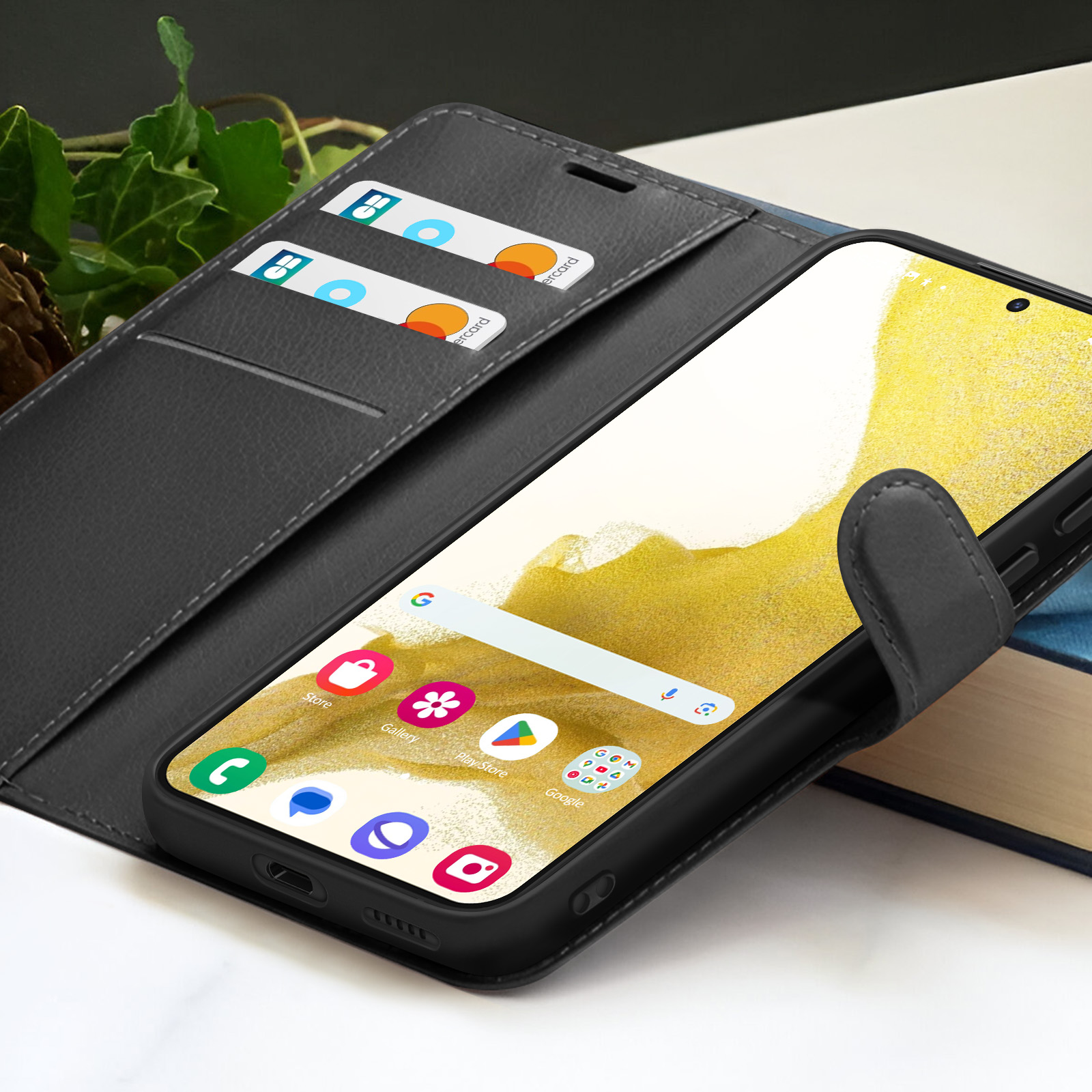 Un smartphone dans un étui noir, ouvert pour révéler l'écran et les fentes pour cartes de crédit. L'arrière-plan comprend des livres et du feuillage.