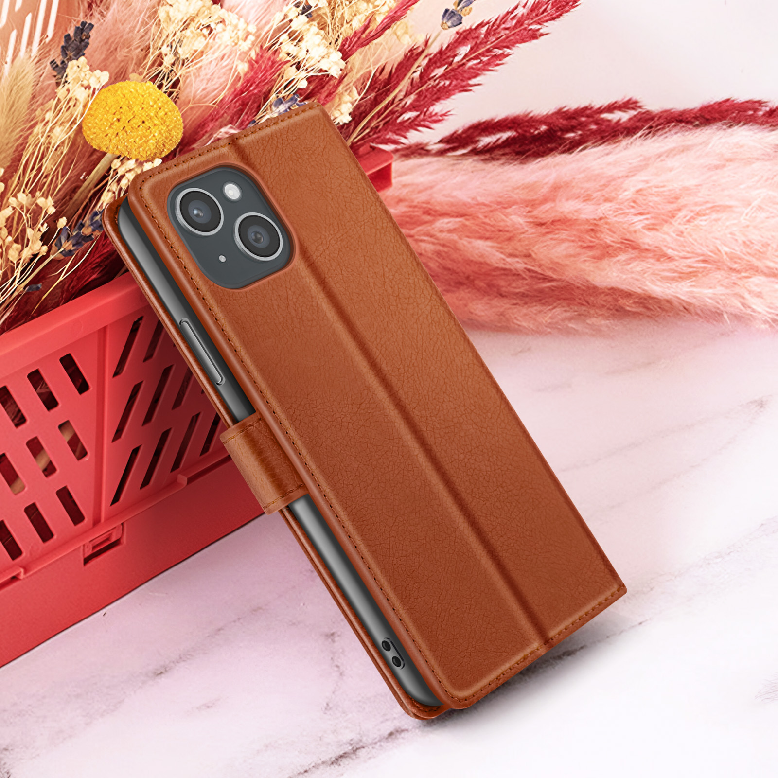 Een bruine leren telefoonhoes met een telefoon erin, rode mand en droogbloemen op een marmeren oppervlak.