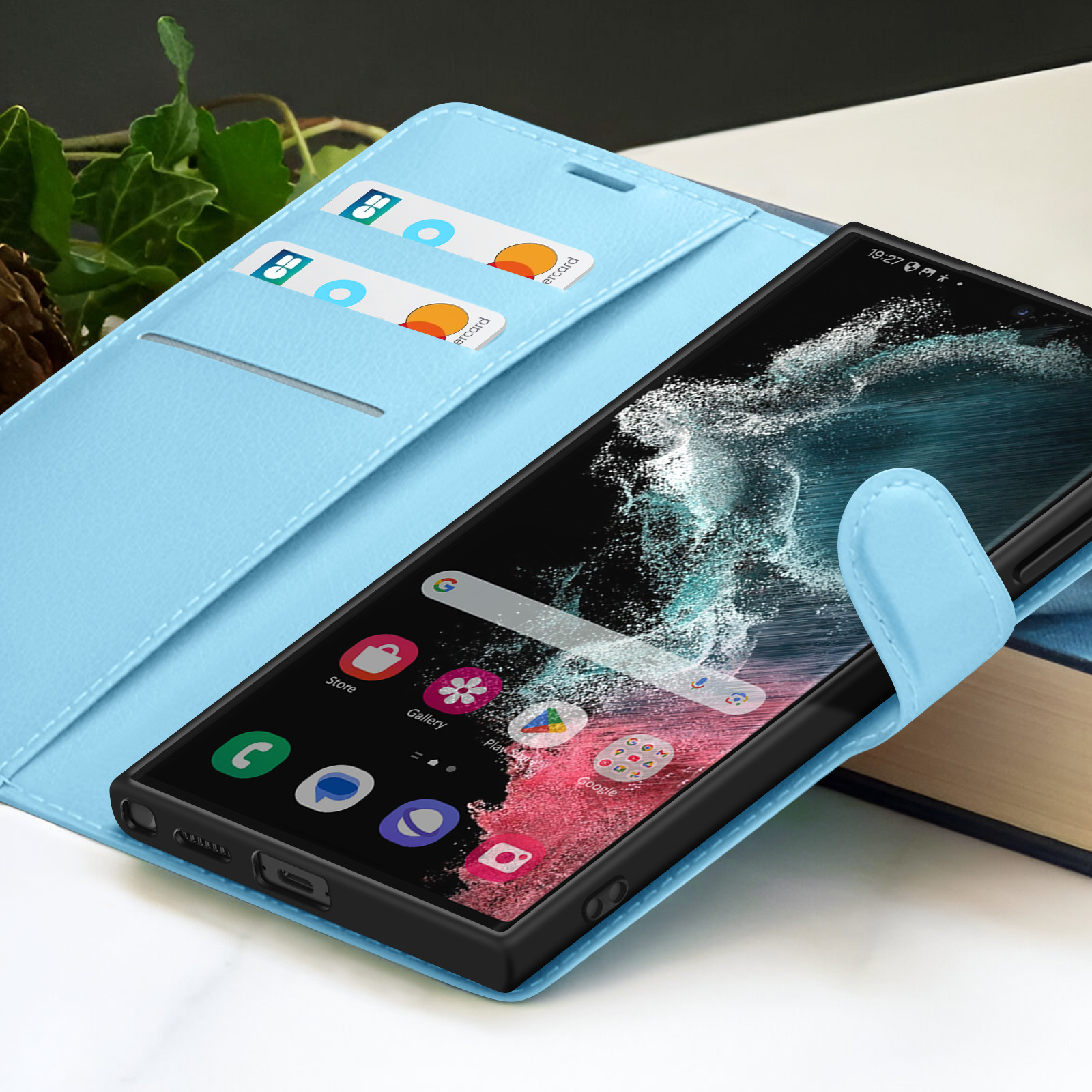 Smartphone dans un étui bleu, affichant des applications. Cartes de crédit dans l'étui. Plante verte en arrière-plan, livres visibles.