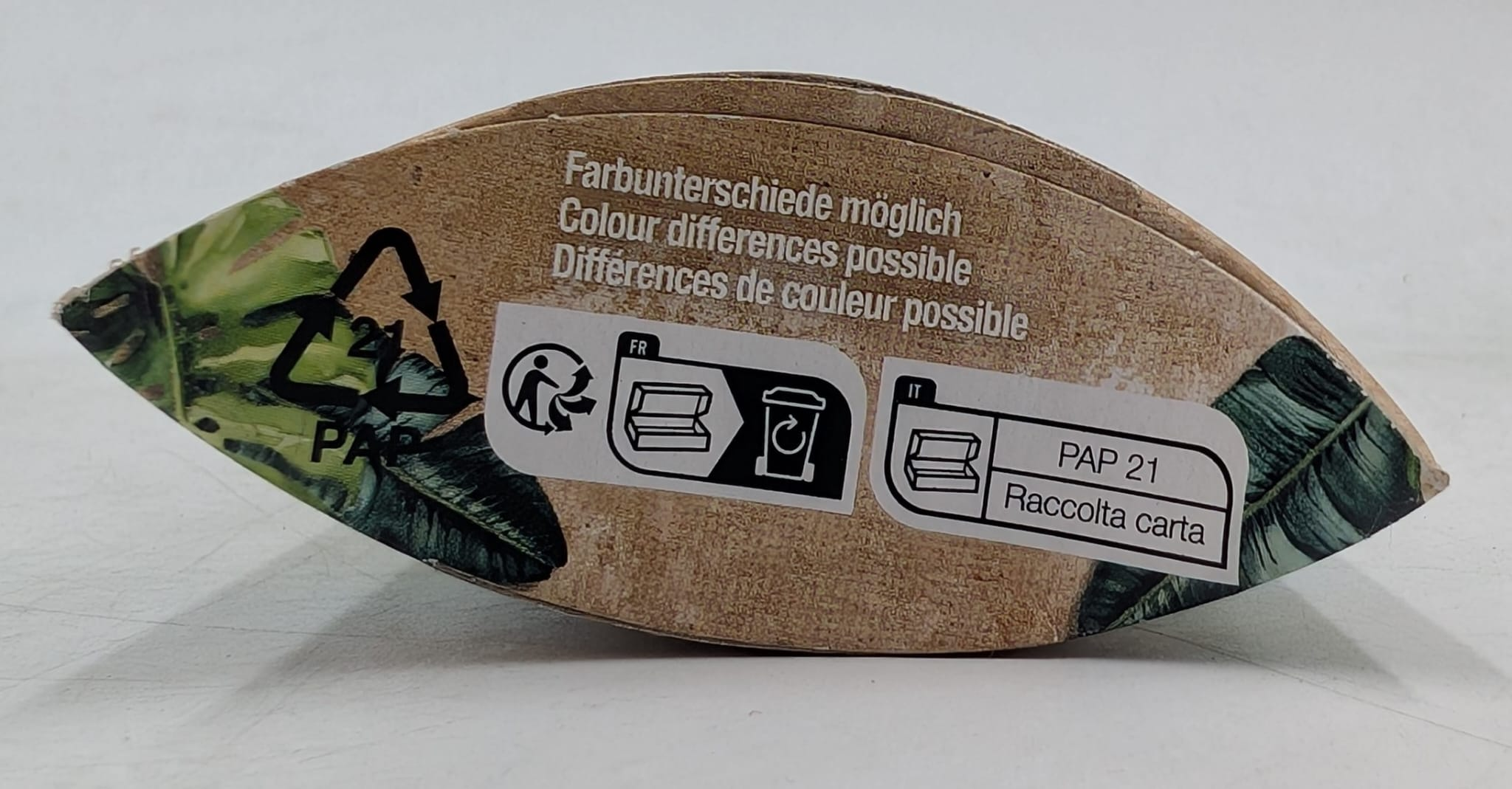 Brązowe papierowe opakowanie z tekstem w wielu językach. Widoczne symbole recyklingu i etykieta PAP 21. Wzór zielonych liści z boku.