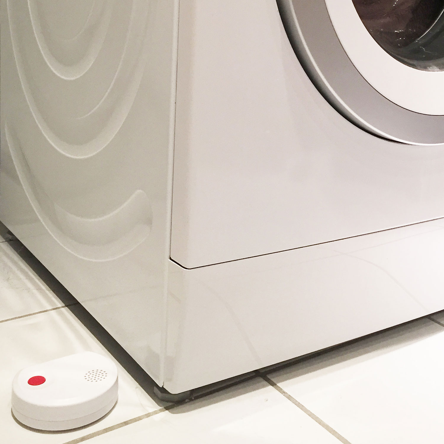 Eine weiße Waschmaschine, ein weißer Sensor mit rotem Knopf und weißer Fliesenboden.