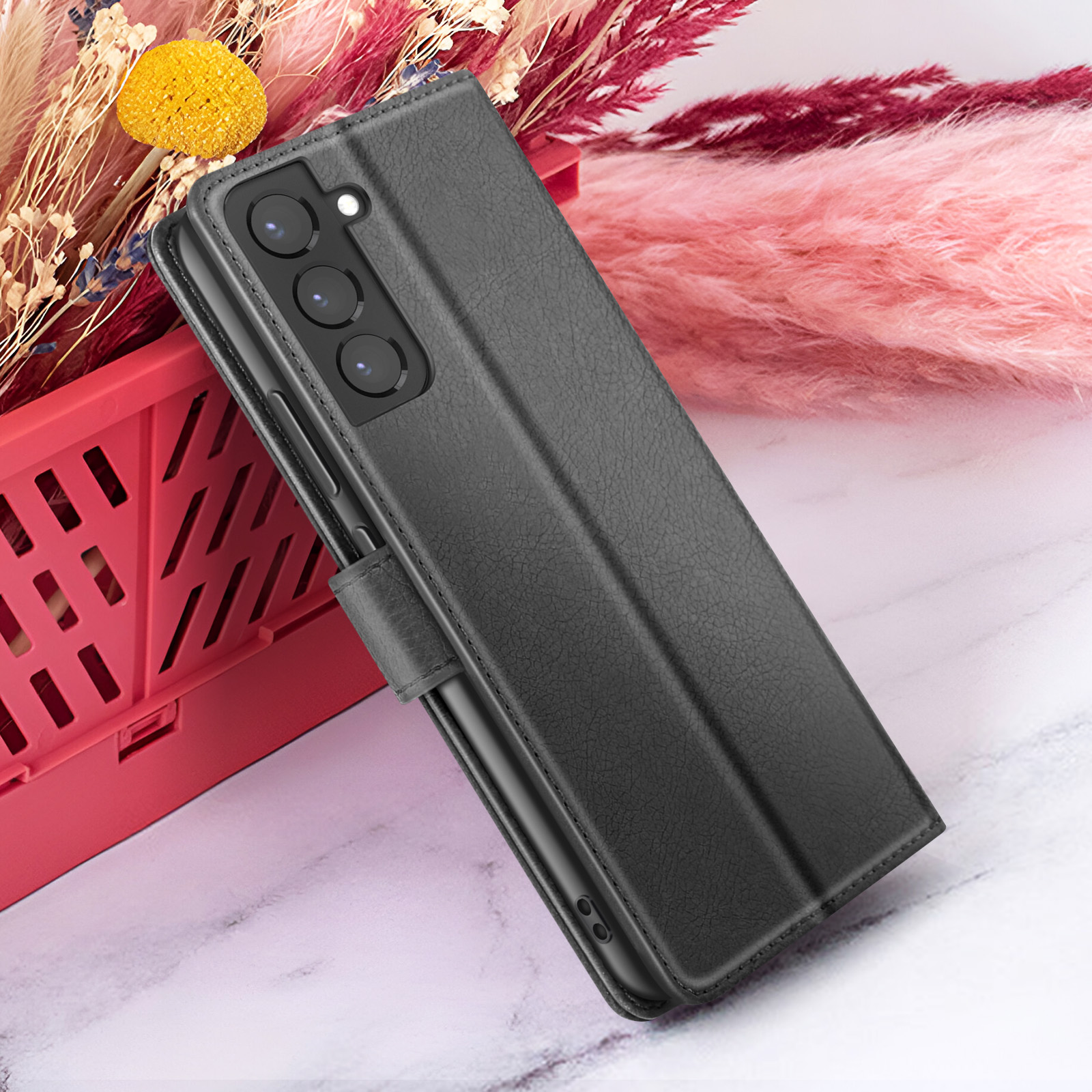 Un étui de téléphone noir avec une surface texturée, ouvert pour révéler un téléphone. À côté, un récipient rouge et des fleurs roses.