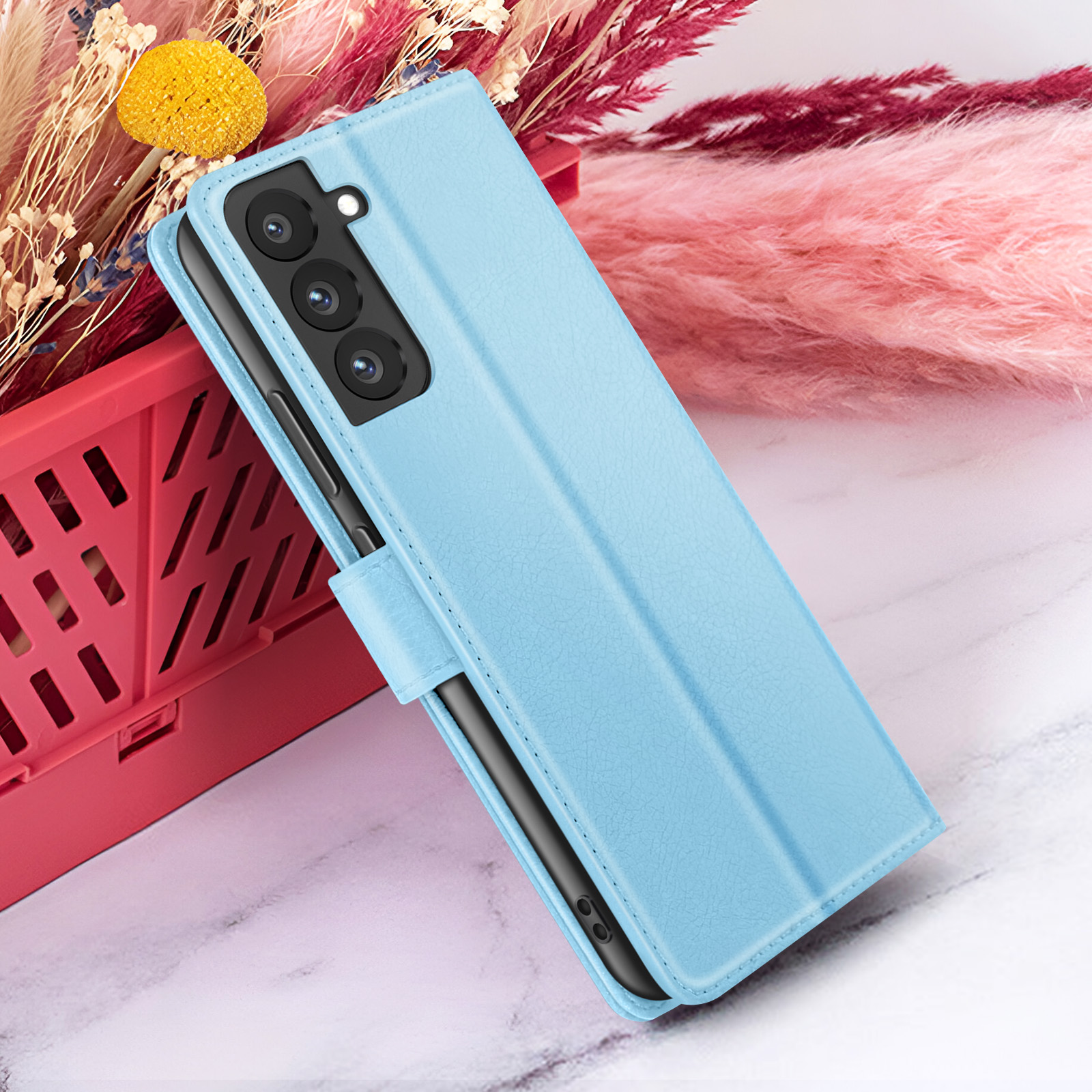 Un étui de téléphone bleu clair avec un téléphone à l'intérieur, placé avec des fleurs roses et un panier rouge sur une surface en marbre blanc.