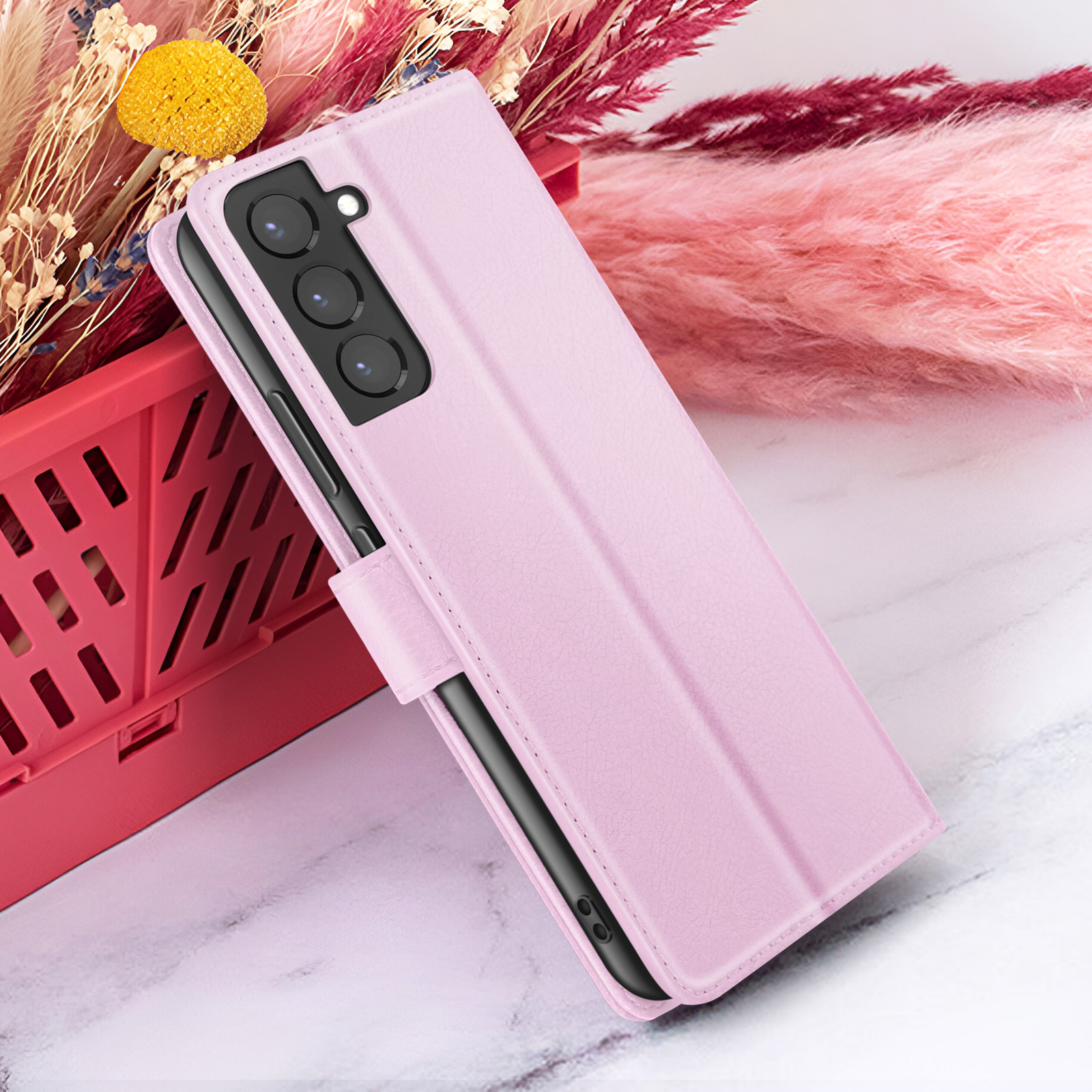 Étui de téléphone rose sur marbre, avec fleurs en arrière-plan. L'étui a une découpe pour la caméra. Panier rouge dans le coin.