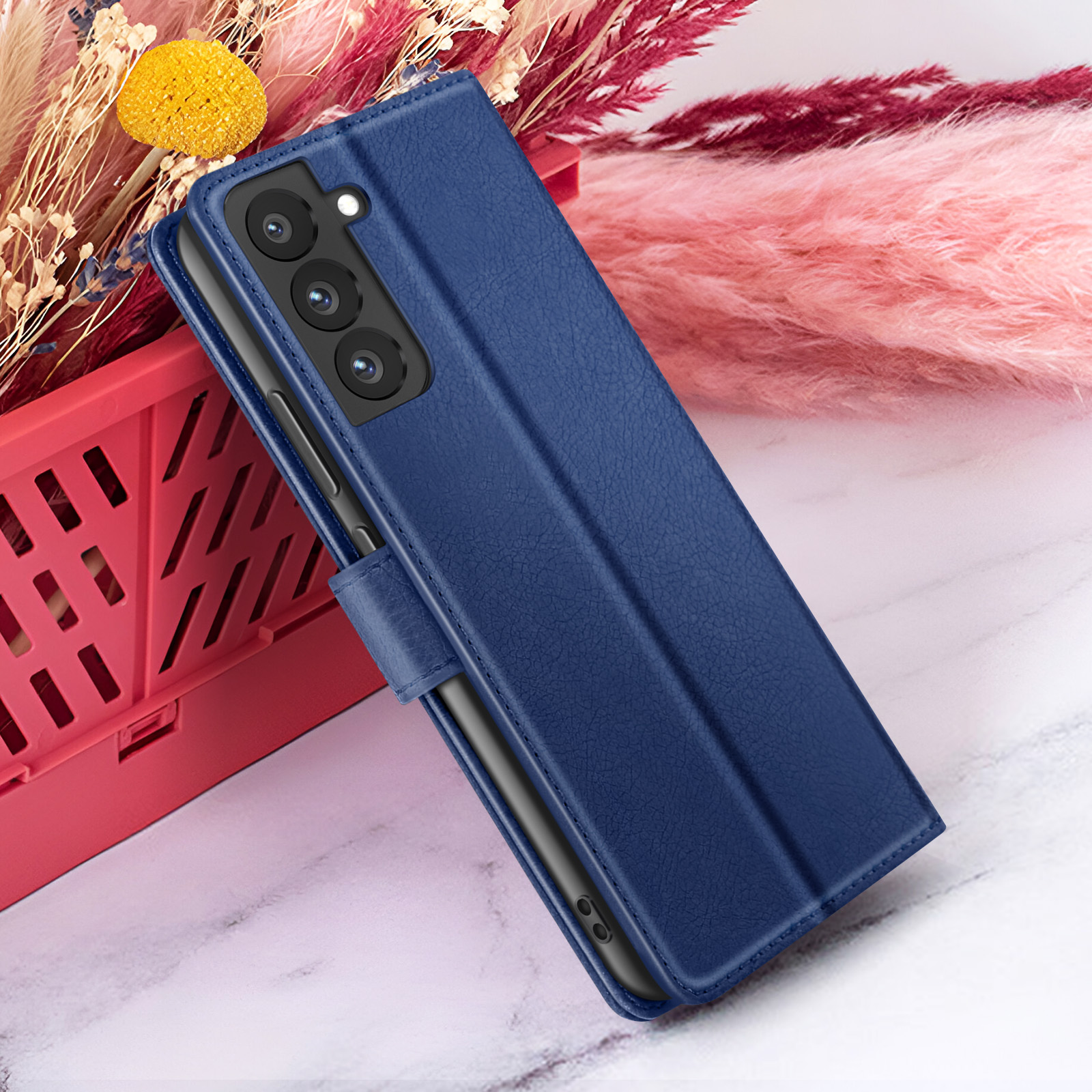 Un étui de téléphone en cuir bleu est sur une surface en marbre, avec des fleurs roses et blanches et un panier rouge en arrière-plan.