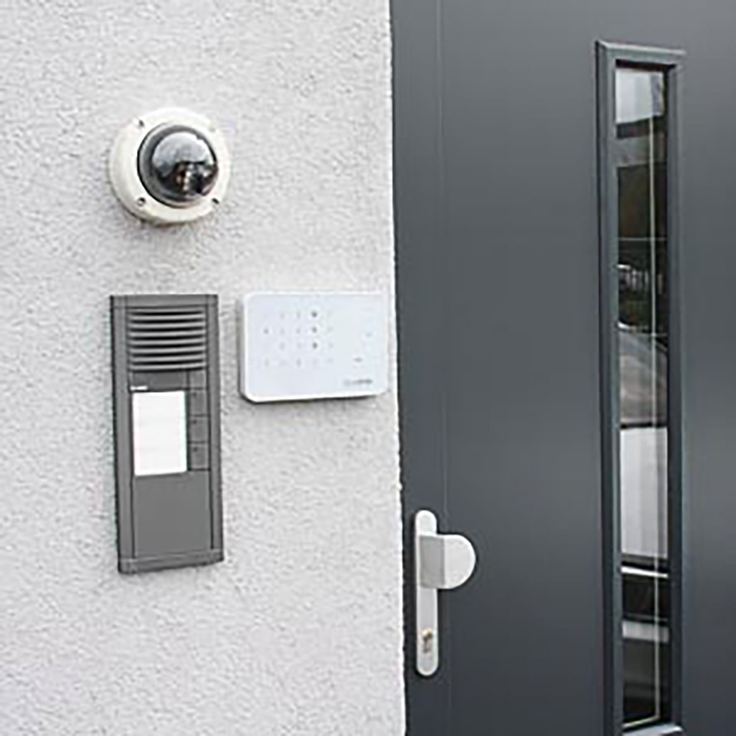 Eine Wand mit Türklingel, Tastatur und Kamera. Rechts eine dunkelgraue Tür mit Fenster und Griff.
