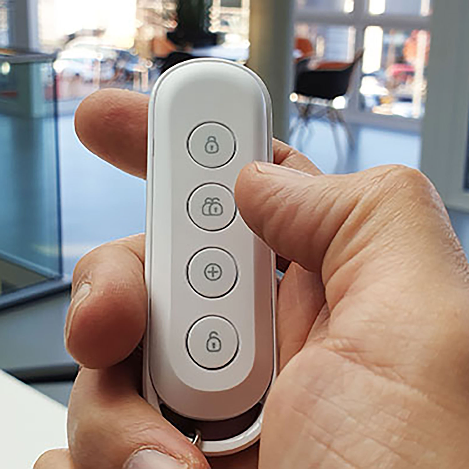 Una mano tiene un telecomando bianco con quattro pulsanti. Lo sfondo mostra un ambiente ufficio sfocato con vetro e mobili.
