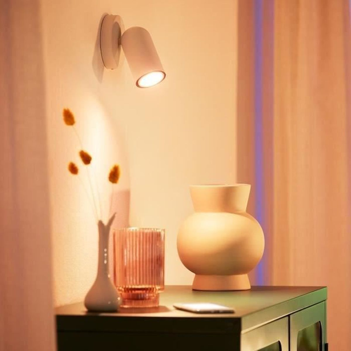 Świecąca lampa ścienna świeci na stół z wazonem, szklanką i suszonymi kwiatami. Na stole spoczywa telefon.