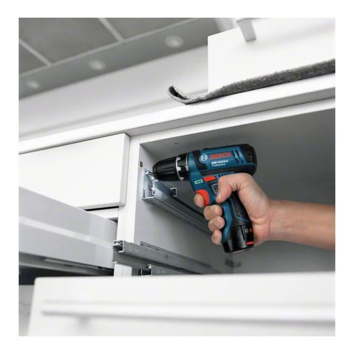 Une main tient une perceuse Bosch bleue et noire, vissant dans un cadre d'armoire blanc. Le fond montre une partie de l'armoire et un objet gris.