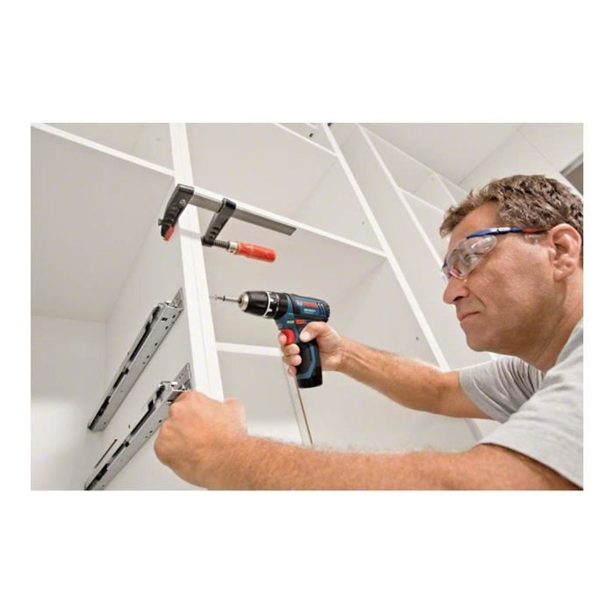 Un homme perce un placard blanc. Il porte des lunettes de sécurité et tient une perceuse. Une pince et des glissières de tiroir sont visibles.