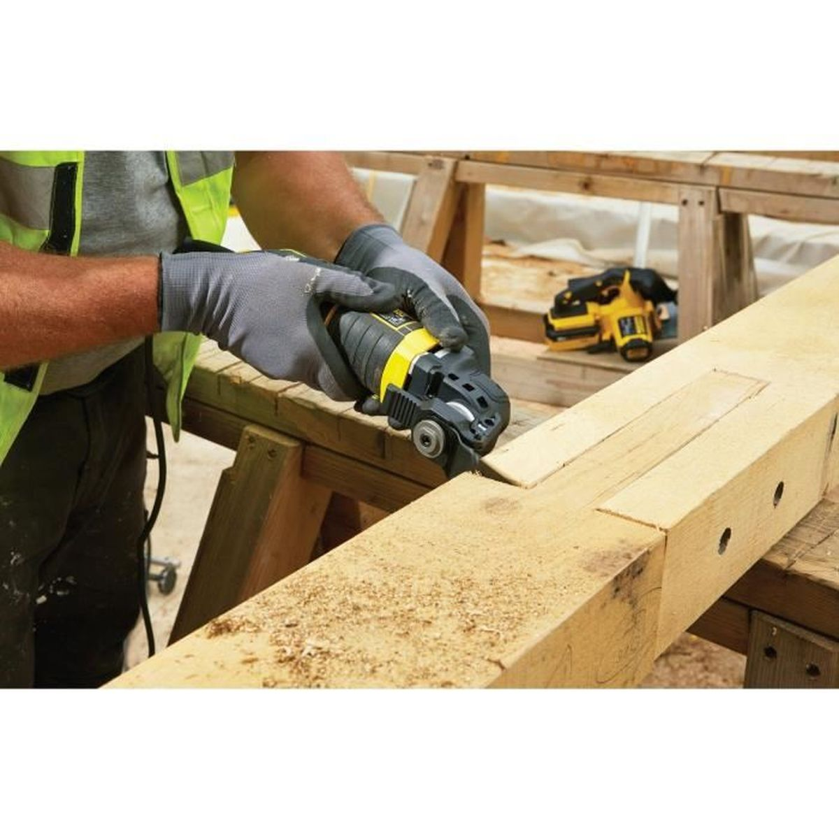 Ein Arbeiter benutzt ein gelb-schwarzes Multitool, um Holz zu schneiden. Ein anderes Werkzeug befindet sich im Hintergrund. Der Arbeiter trägt Handschuhe und eine Sicherheitsweste.
