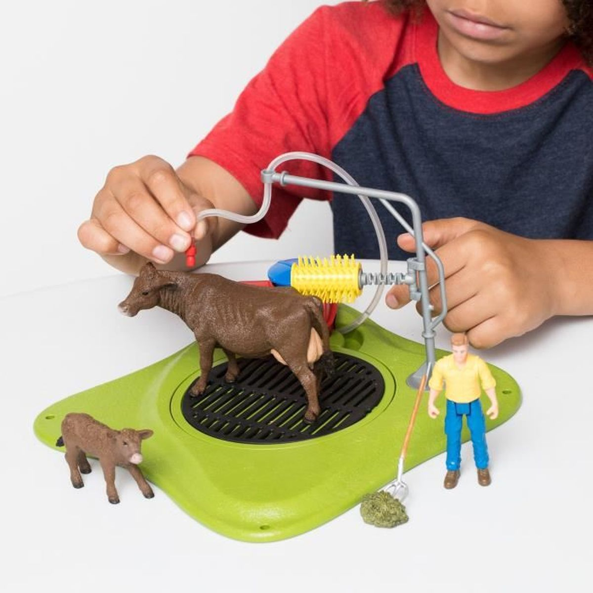 Ein Kind spielt mit einem Spielzeug-Bauernhof-Set mit einer Kuh, einem Kalb, einem Bauern und einer Reinigungsstation auf einer grünen Plattform.