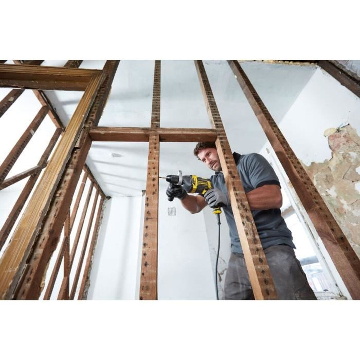Hombre taladrando un marco de madera con un taladro amarillo y negro. La habitación está en construcción, con vigas de madera expuestas.