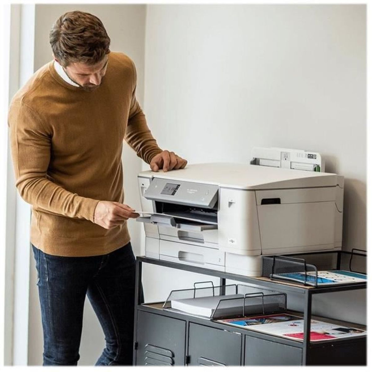 Mężczyzna wkłada papier do dużej drukarki. Ma na sobie brązowy sweter i dżinsy. Drukarka na szafce z tacami na papier.