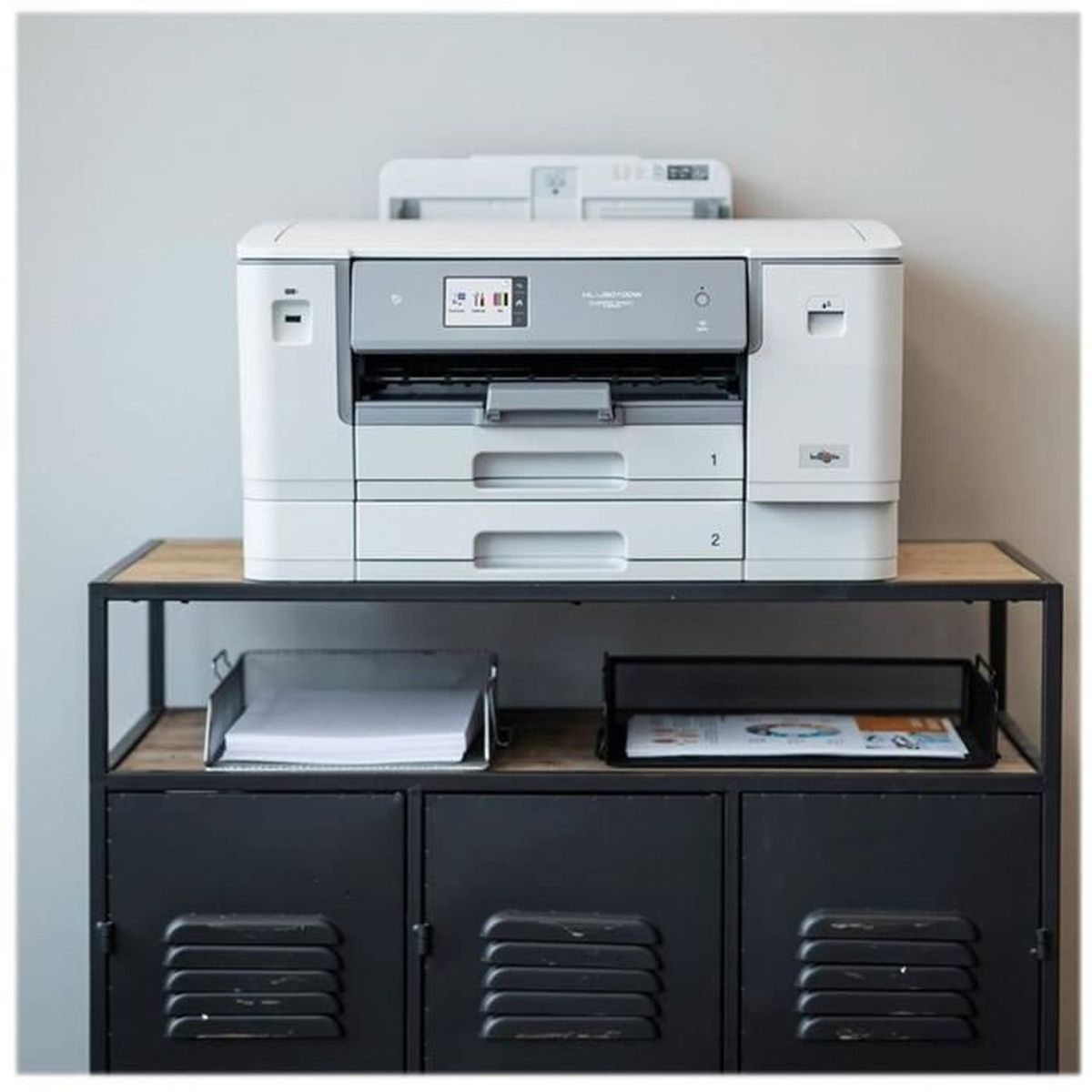 Biała drukarka stoi na czarnej szafce. Poniżej tace na papier. Ściana jest szara.