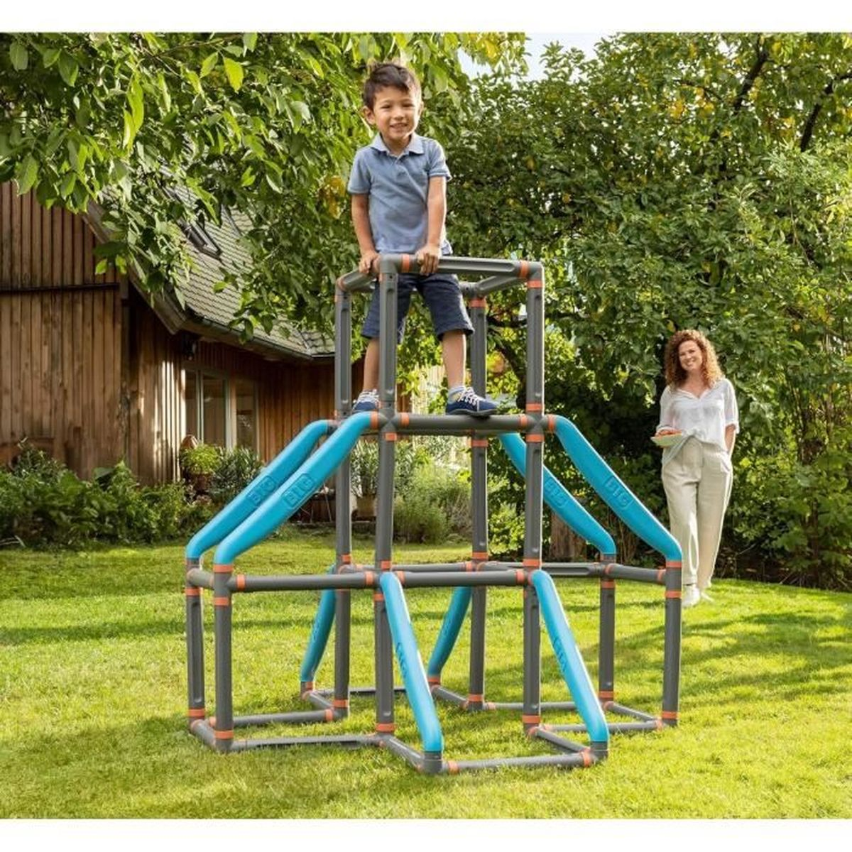 Ein Junge steht auf einem Klettergerüst. Im Hintergrund steht eine Frau. Die Szene spielt in einem grasbewachsenen Garten mit Bäumen und einem Haus.