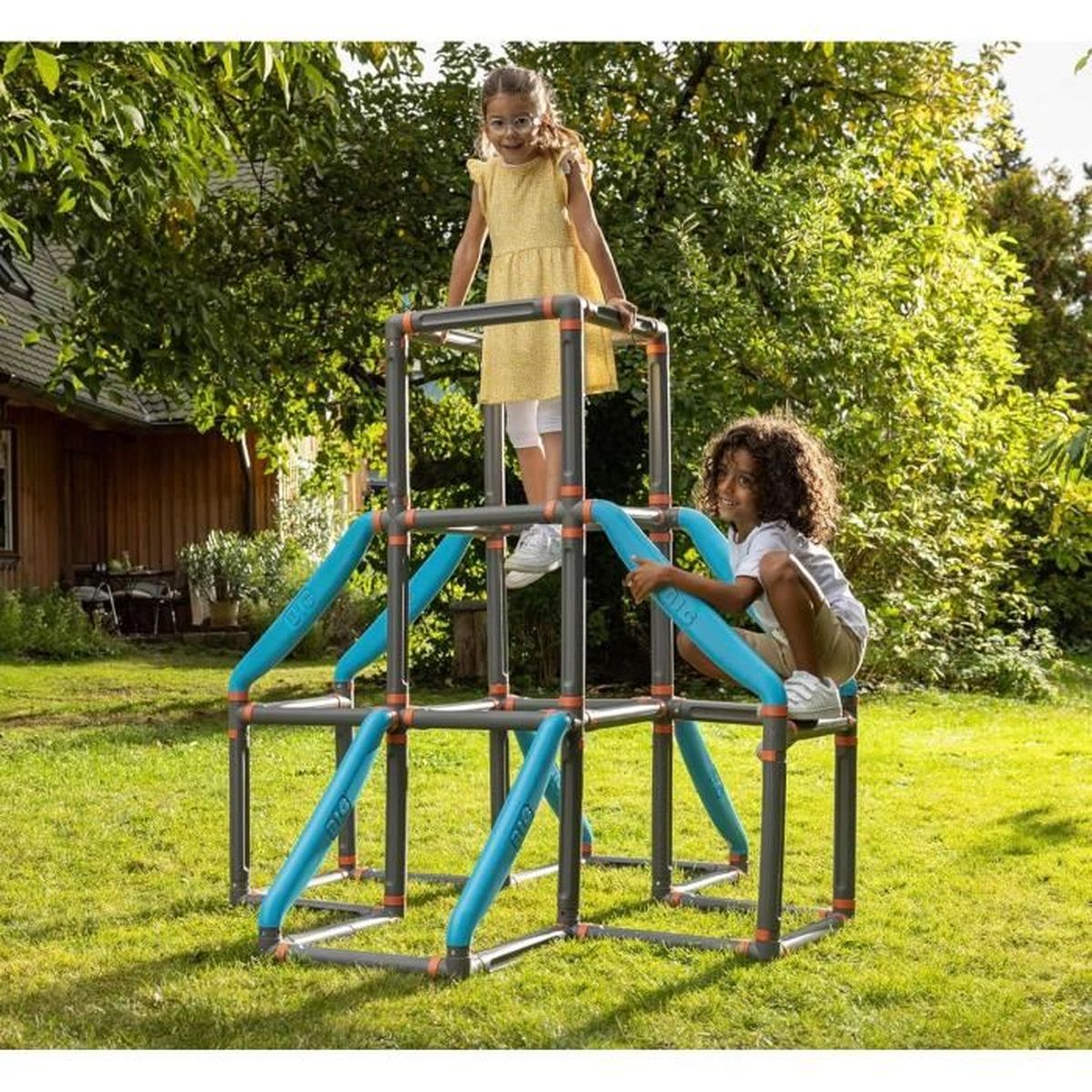 Zwei Kinder spielen auf einem grau-blauen Klettergerüst in einem grasbewachsenen Hinterhof. Ein Kind ist oben, das andere klettert.