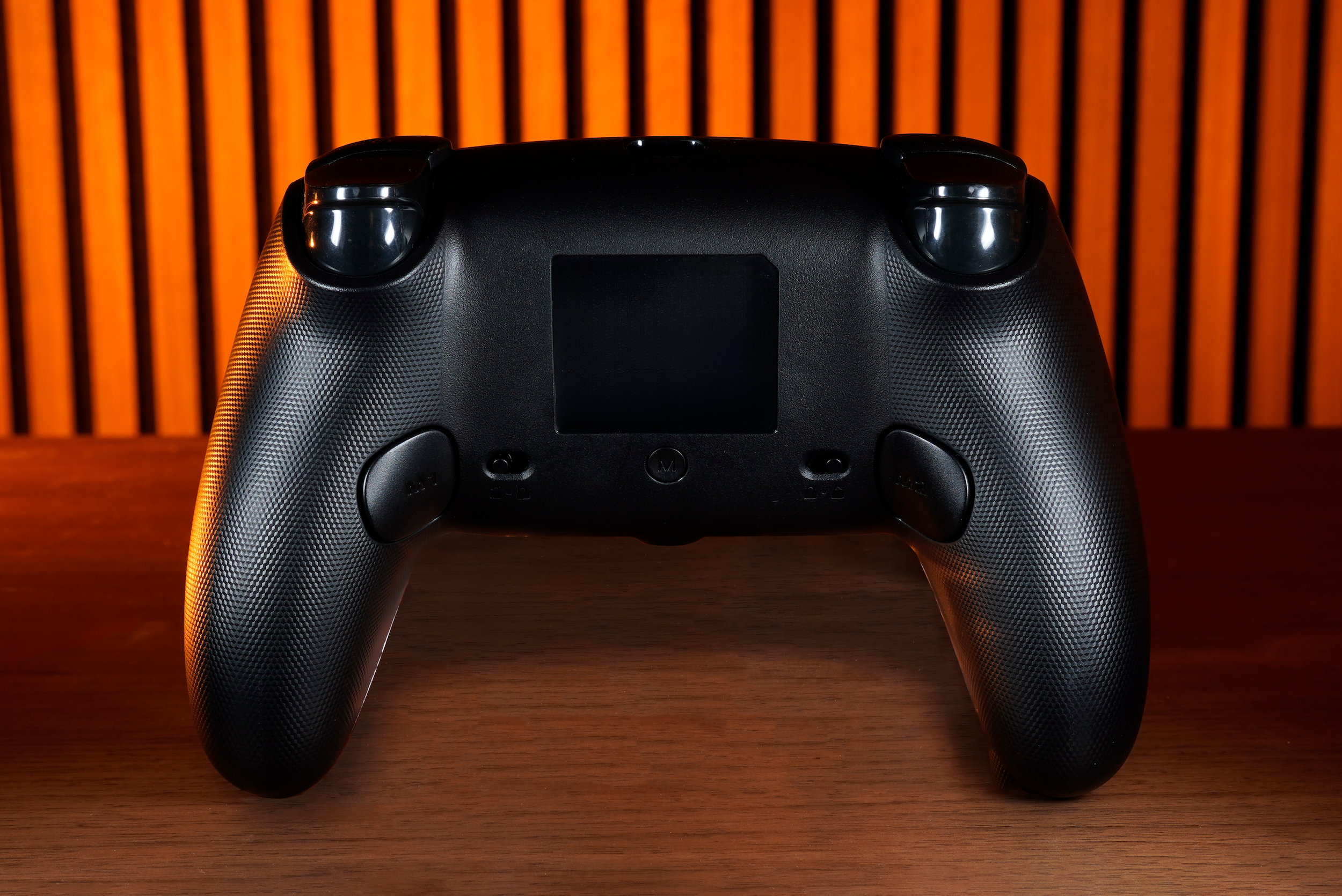 A black game controller sits on a wooden surface. The background is orange with black vertical stripes.