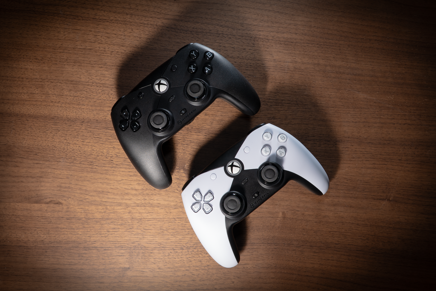 Two Xbox controllers, one black and one white, on a wooden surface. Both controllers are facing up.