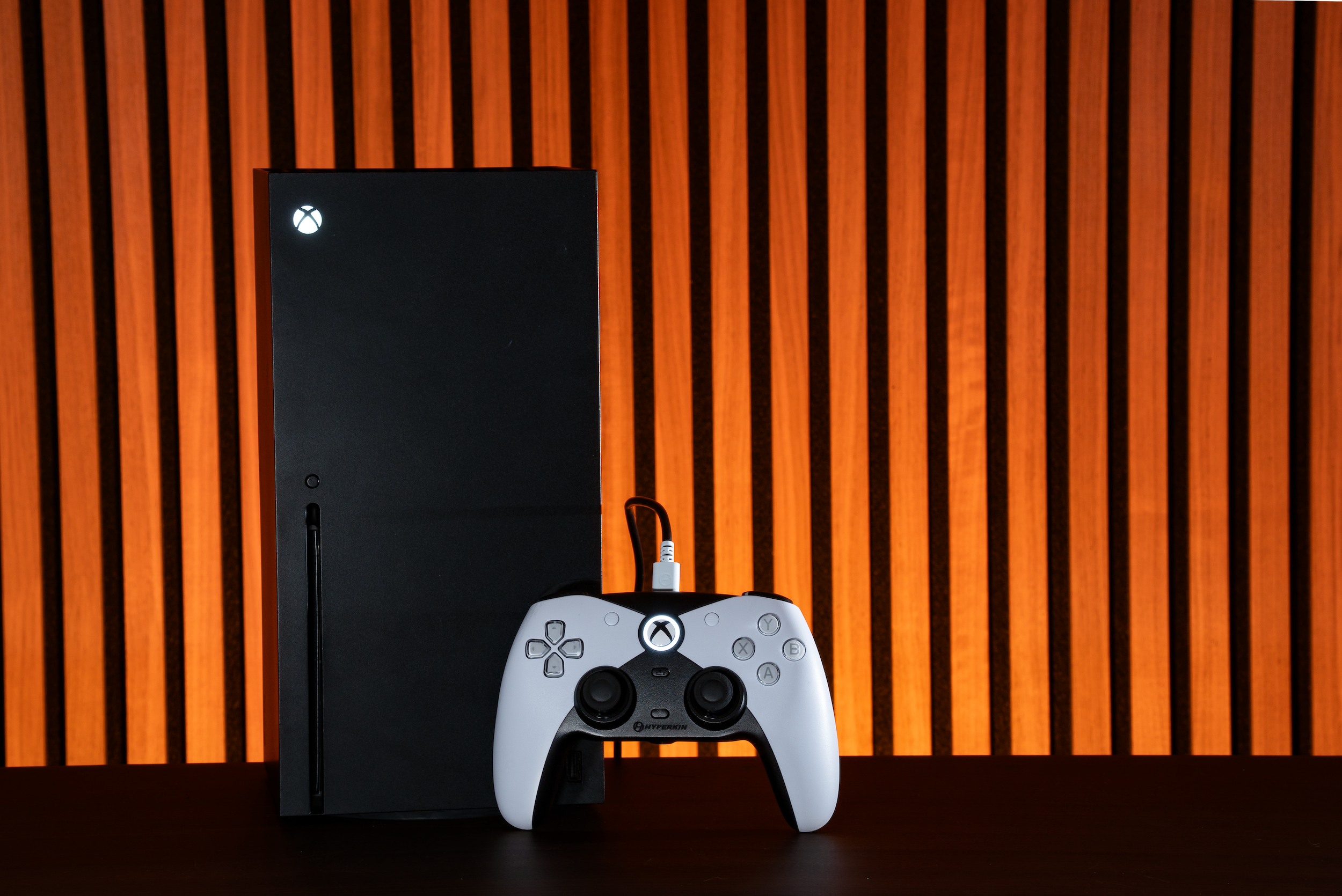 Una console Xbox Series X nera accanto a un controller bianco e nero. Lo sfondo è un pannello di legno arancione.