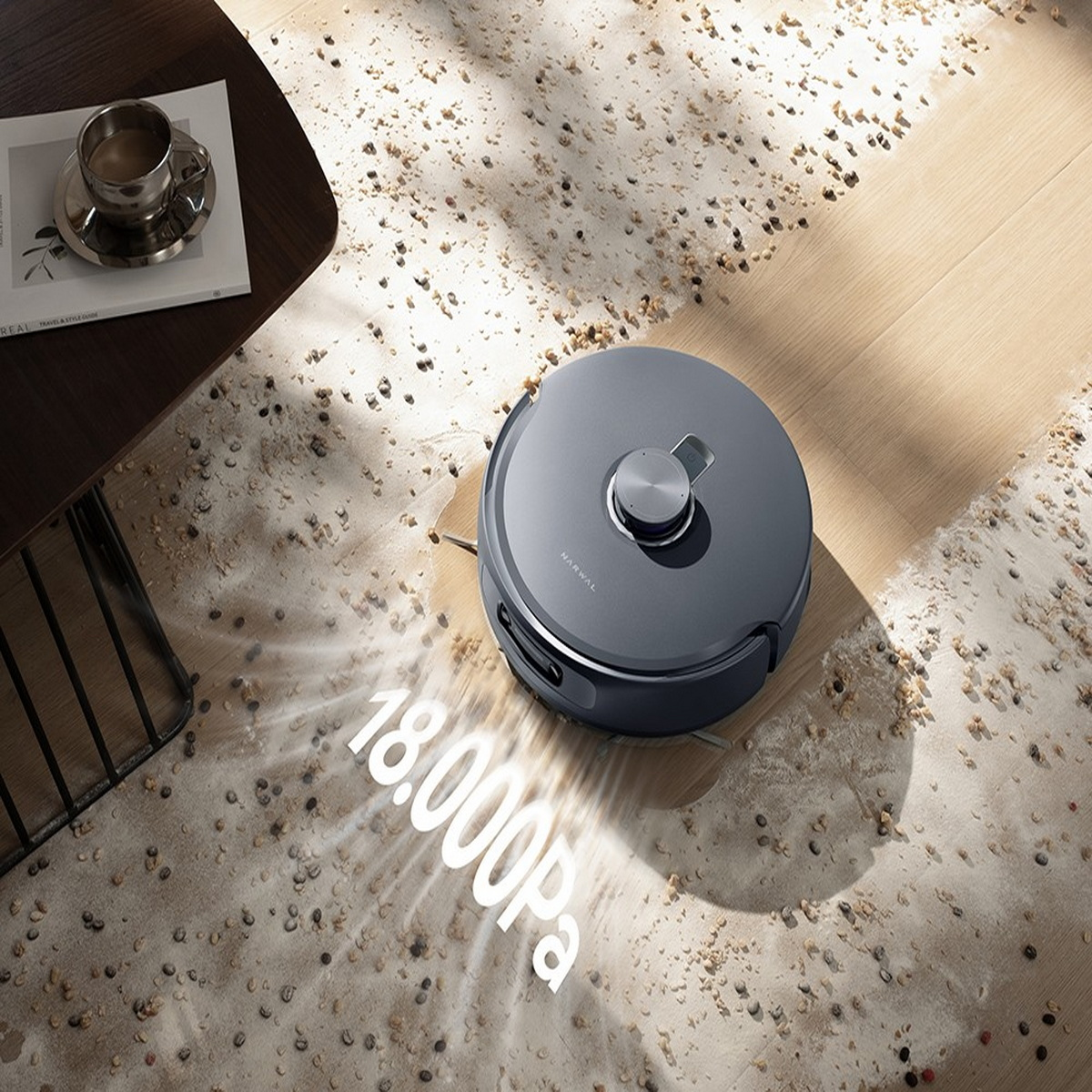Ein grauer Roboter-Staubsauger reinigt auf einem Holzboden Schmutz. Eine Kaffeetasse und ein Buch stehen auf einem Tisch. Text: 18.000Pa.
