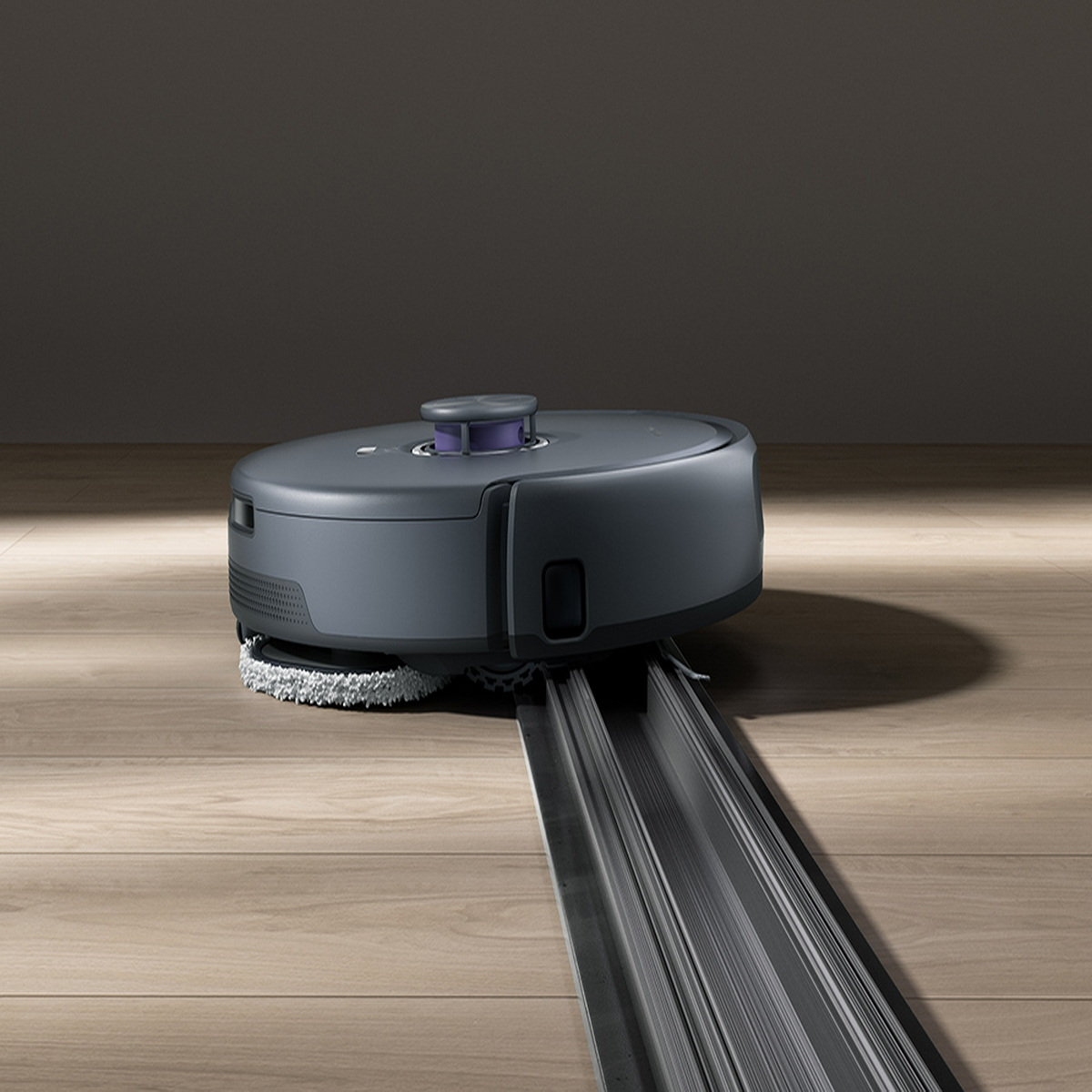 Ein dunkelgrauer Roboter-Staubsauger auf einem Holzboden mit einer Schiene. Er hat einen lila Deckel und ein weißes Reinigungspad.