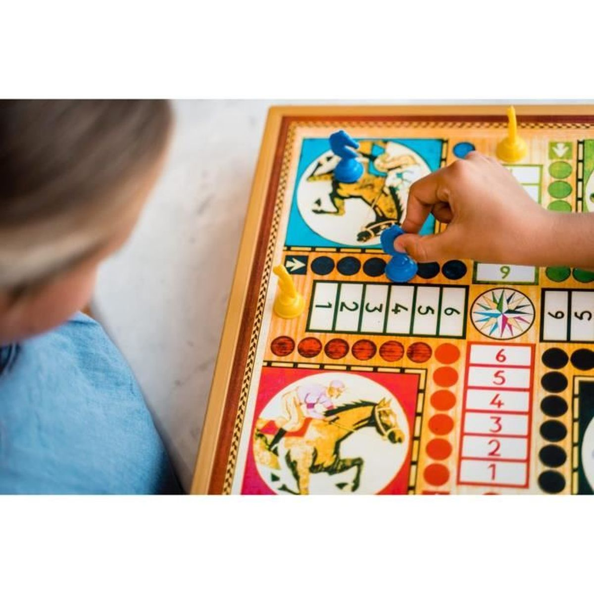 Die Hand einer Person platziert ein blaues Spielteil auf einem Pferderennspiel. Das Brett ist bunt mit Zahlen und Pferden.