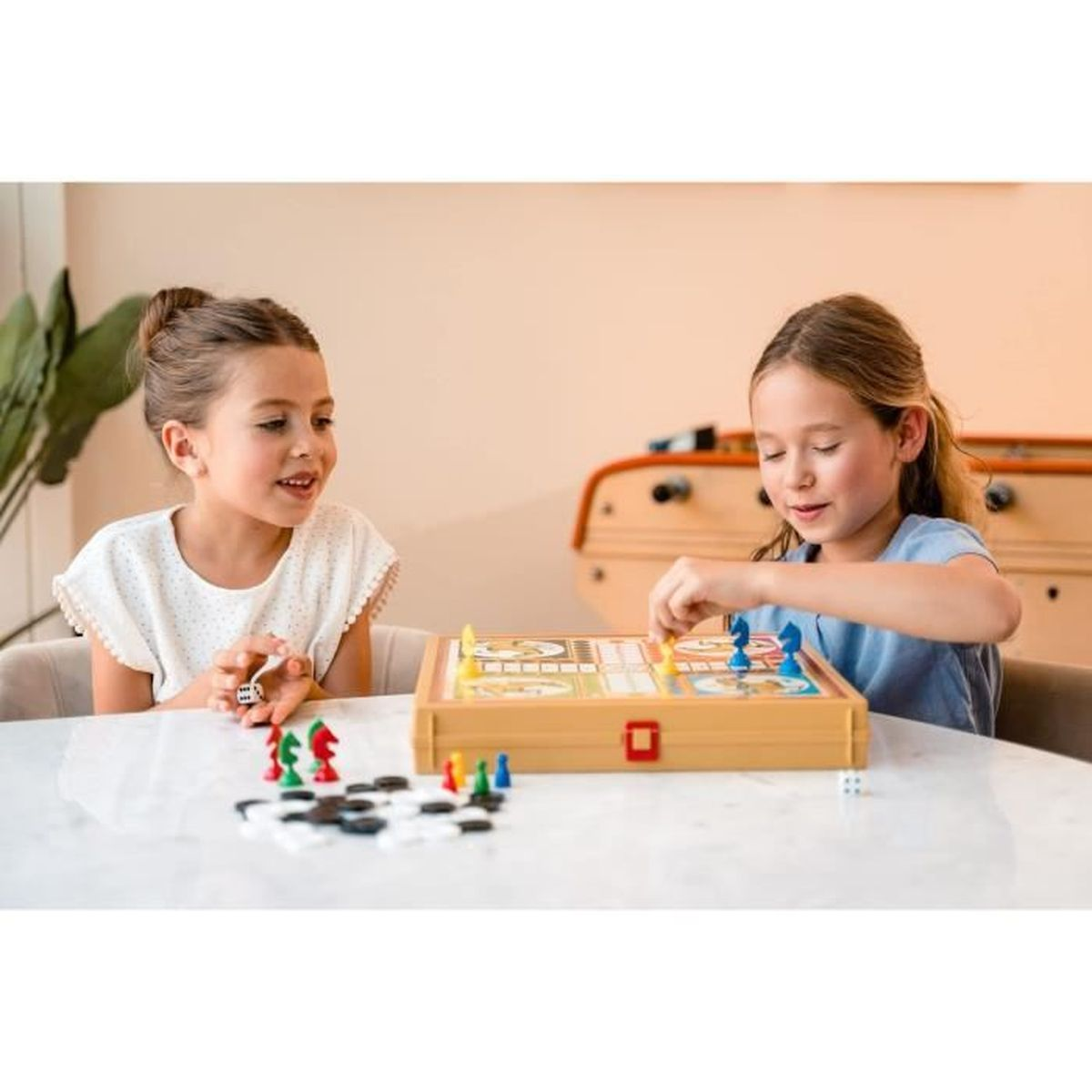 Zwei junge Mädchen spielen ein Brettspiel. Eines hält Würfel. Spielbrett, Figuren und Würfel auf weißem Tisch. Beigefarbene Wand im Hintergrund.