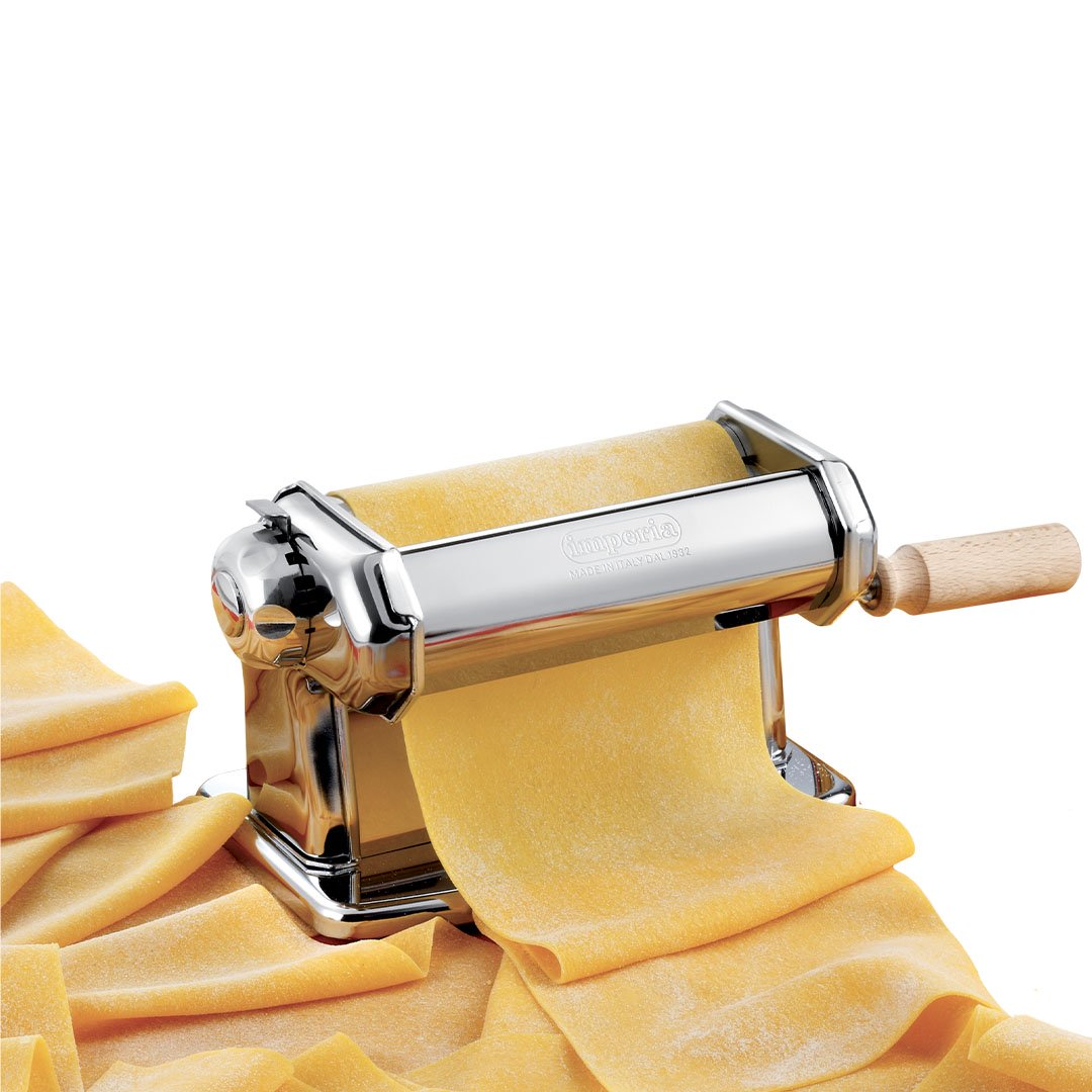 Una macchina per la pasta con manico in legno sta facendo pasta fresca. Escono fogli di pasta gialla. Altri fogli di pasta sono in primo piano.
