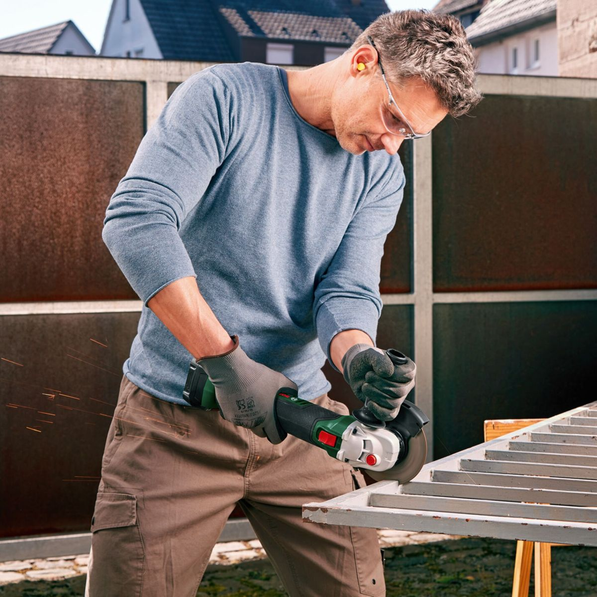 Mężczyzna używa szlifierki kątowej. Ma okulary i zatyczki do uszu, tnie metalową ramę. Iskry lecą. Tło: płot i domy.