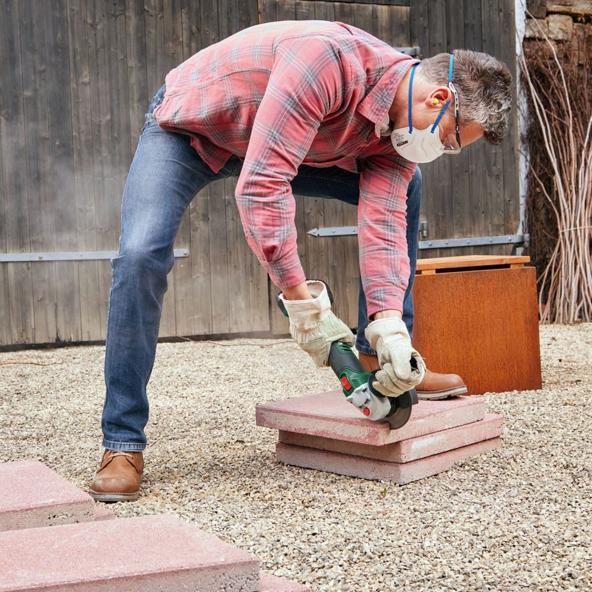 Mężczyzna tnie czerwoną cegłę szlifierką. Nosi okulary ochronne, maskę i rękawice. Tło: drewniane drzwi i żwirowa ziemia.