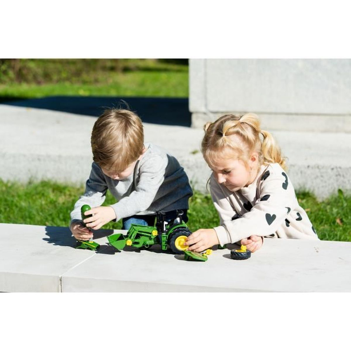 Due bambini giocano con un trattore giocattolo verde su una superficie di cemento. Uno tiene in mano un oggetto verde, l'altro lavora su una ruota.