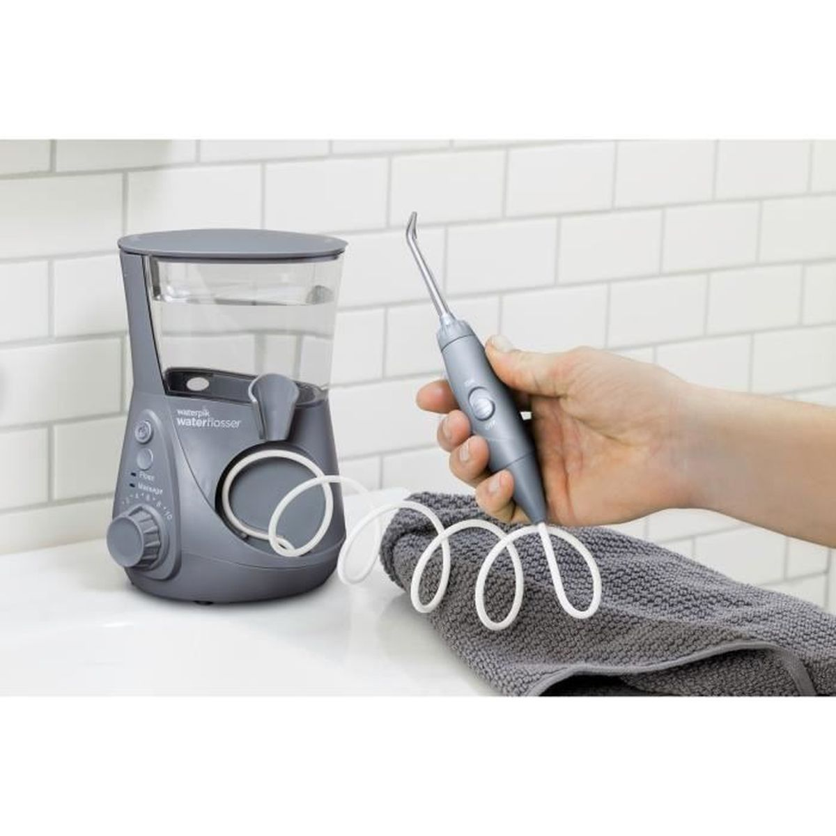 Ein grauer Waterpik-Wasserflosser, mit einer Hand, die den Flosserkopf hält. Ein Handtuch liegt davor, und eine weiß geflieste Wand dahinter.
