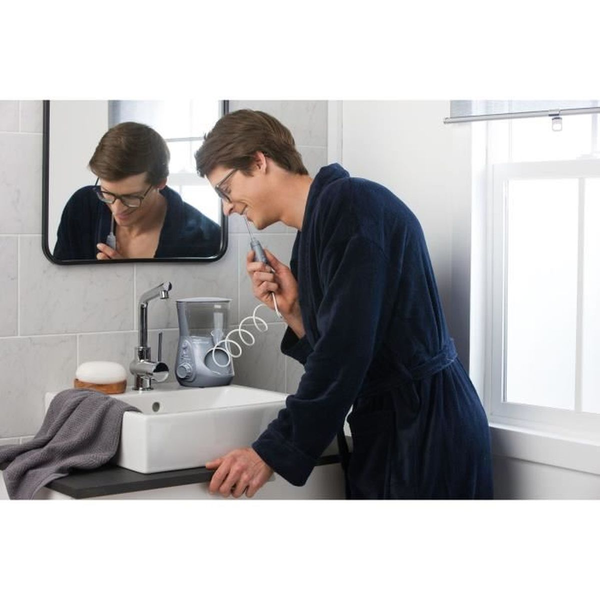 Ein Mann im blauen Bademantel reinigt seine Zähne. Ein Spiegel zeigt sein Spiegelbild. Ein Wasserflosser und ein Waschbecken sind sichtbar.