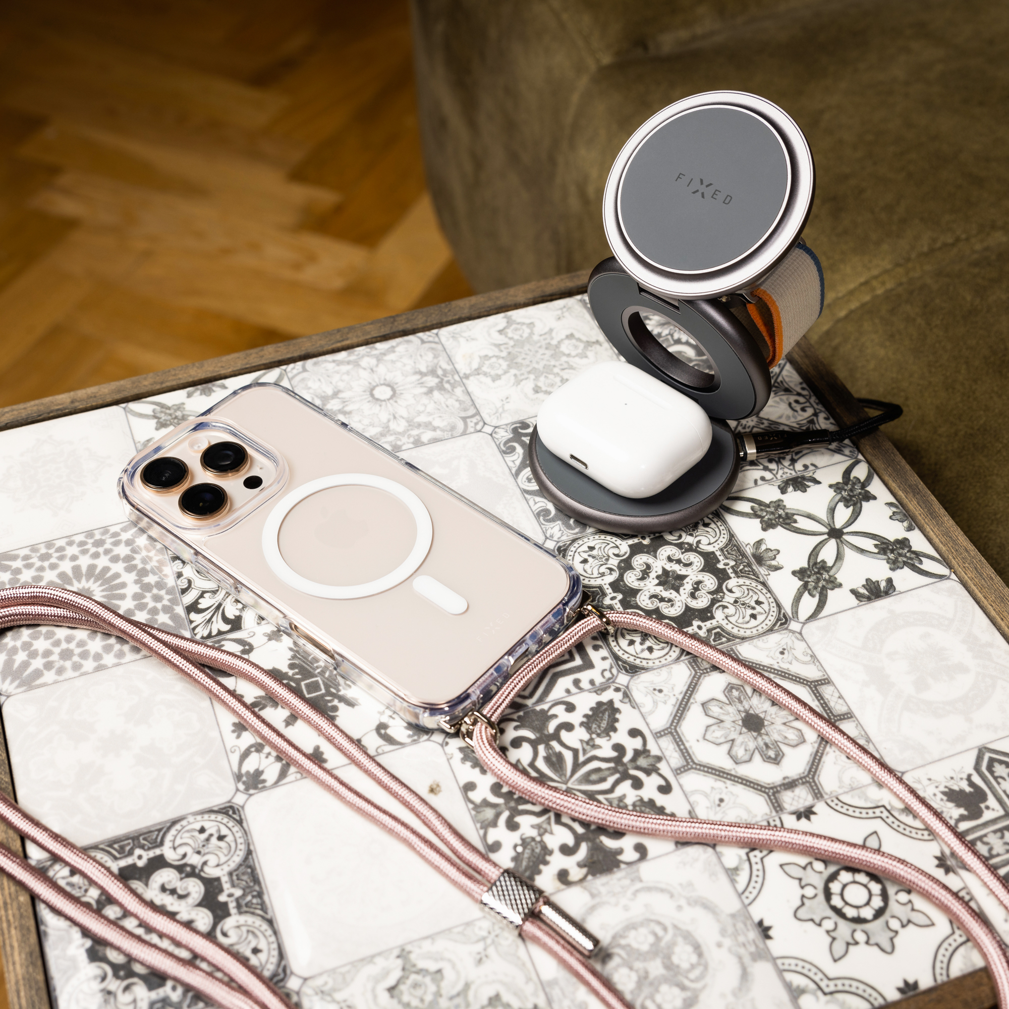 Ein Telefon mit transparentem Gehäuse und rosa Gurt, AirPods und ein kabelloses Ladegerät auf einer gemusterten Oberfläche.