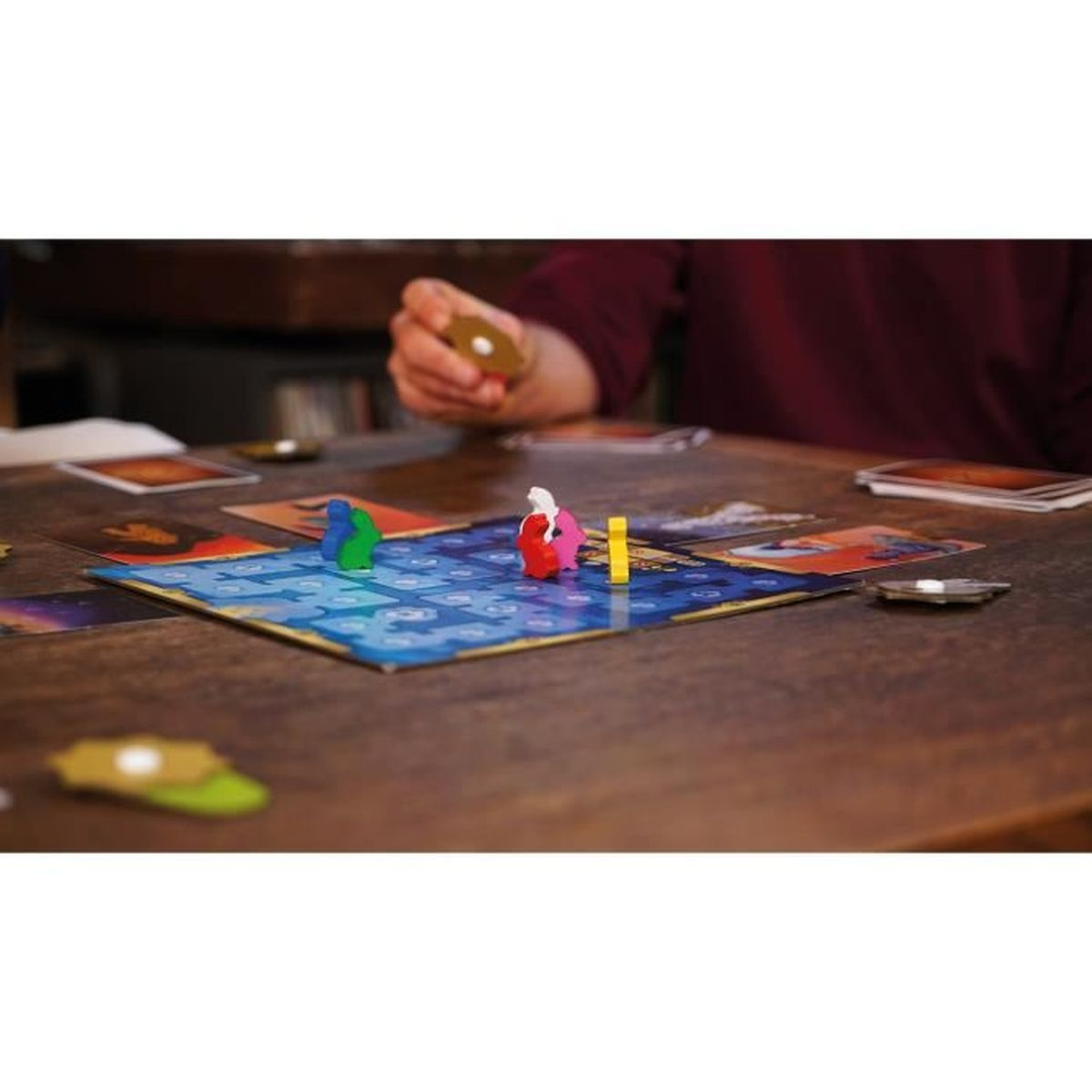 Brettspiel im Gange. Figuren auf einem blauen Brett. Karten und eine Hand, die ein Token hält, sind auf einem Holztisch zu sehen.