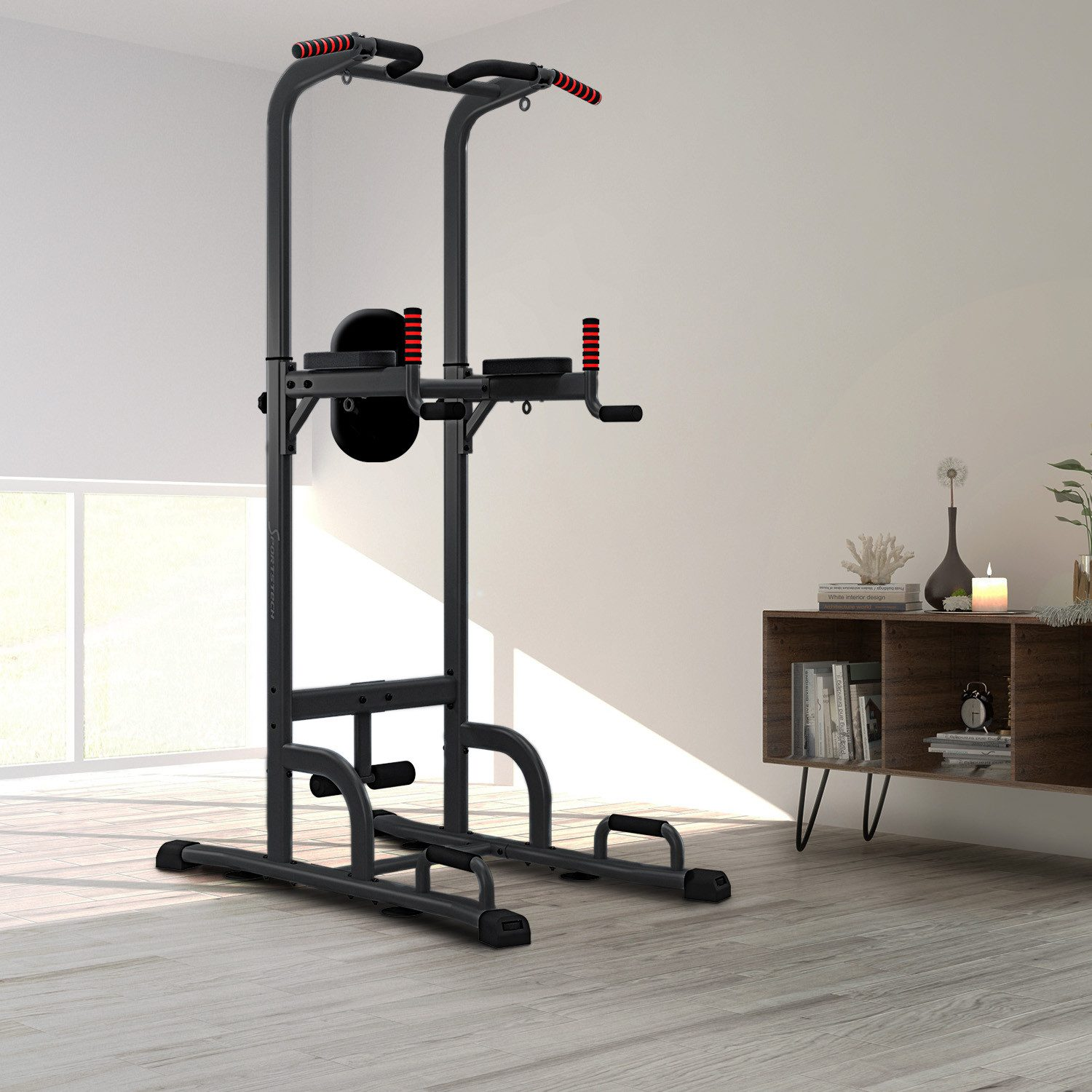 Eine schwarze und rote Trainingsstation steht in einem Raum. Im Hintergrund befinden sich ein Holzschrank und ein Fenster. Der Boden ist helles Holz.