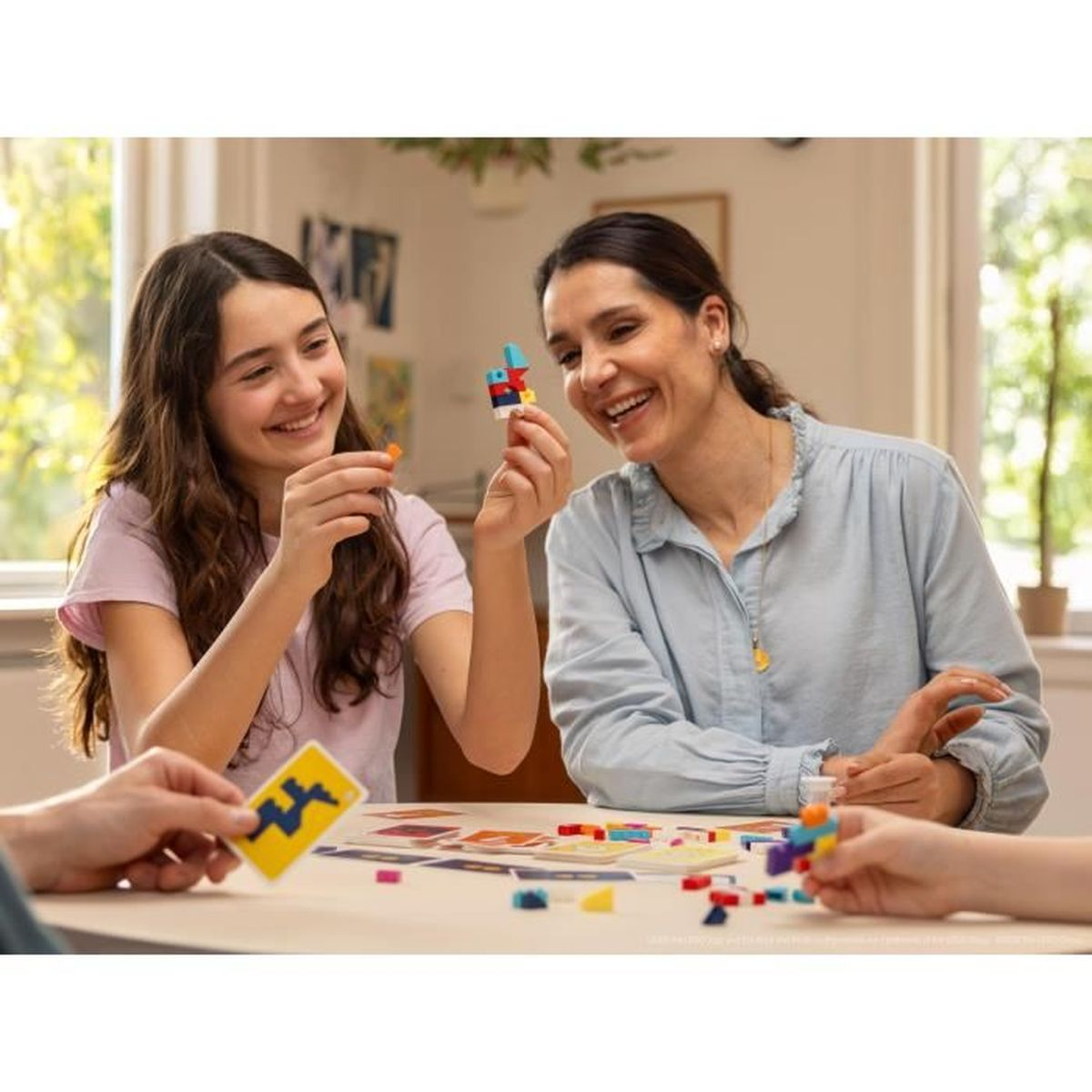Mutter und Tochter spielen ein Bausteinspiel und lächeln. Blöcke und Karten liegen auf dem Tisch. Im Hintergrund ist ein Fenster mit grünen Bäumen.