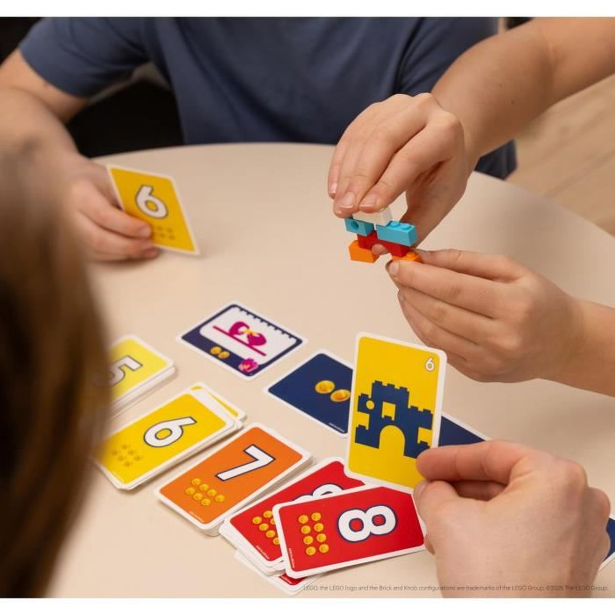 Hände halten bunte Blöcke und Karten mit Zahlen und Bildern auf einem Tisch. Spielkarten sind verstreut.