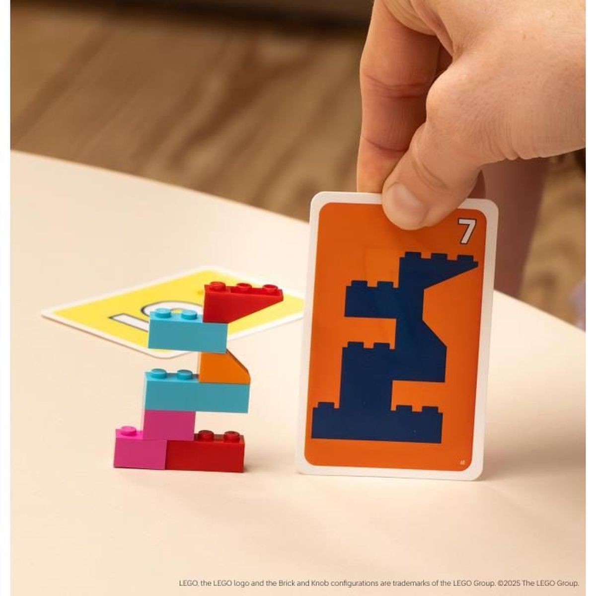 Eine Hand hält eine Karte mit einer blauen Form und der Zahl 7. Bunte LEGO-Steine ​​liegen neben einer gelben Karte.