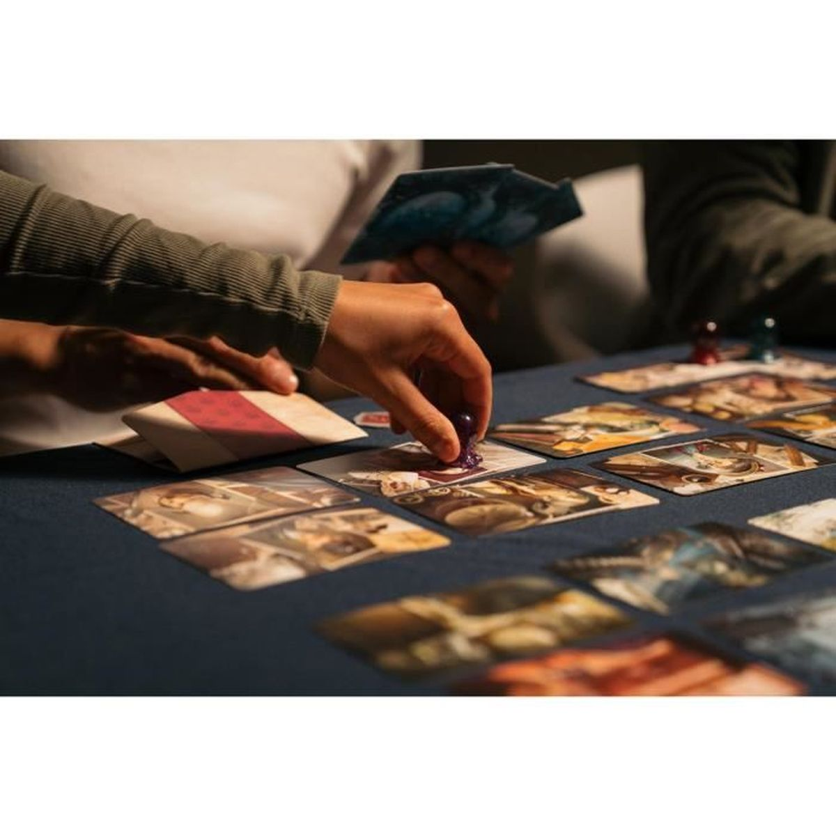 Hände spielen ein Brettspiel mit Karten und Spielsteinen auf einer blauen Oberfläche. Eine Hand legt einen lila Spielstein auf eine Karte.